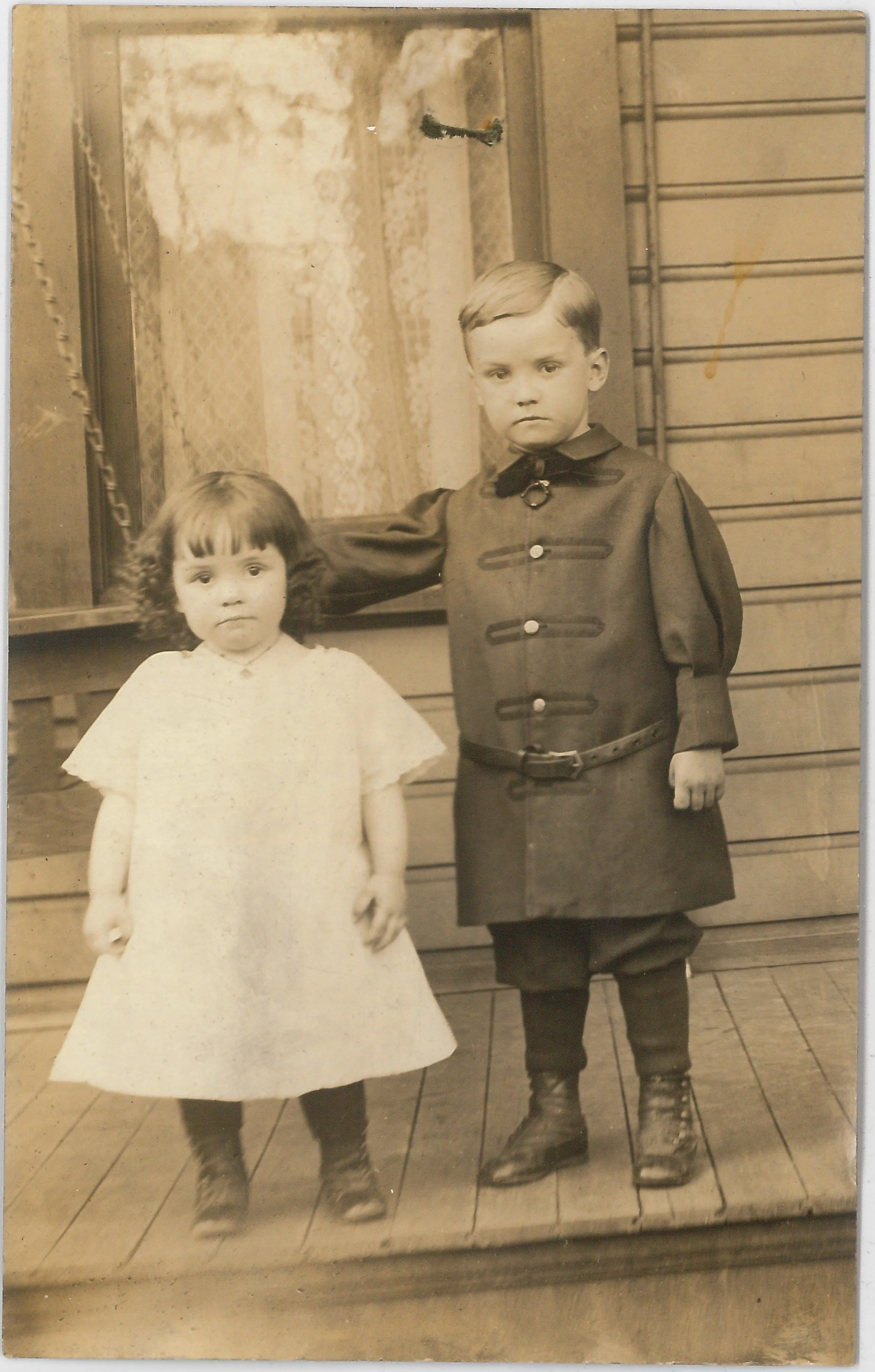 A vintage black and white photo of a young boy and girl standing outdoors on a wooden porch, the boy in uniform and the girl in a light-colored dress.
