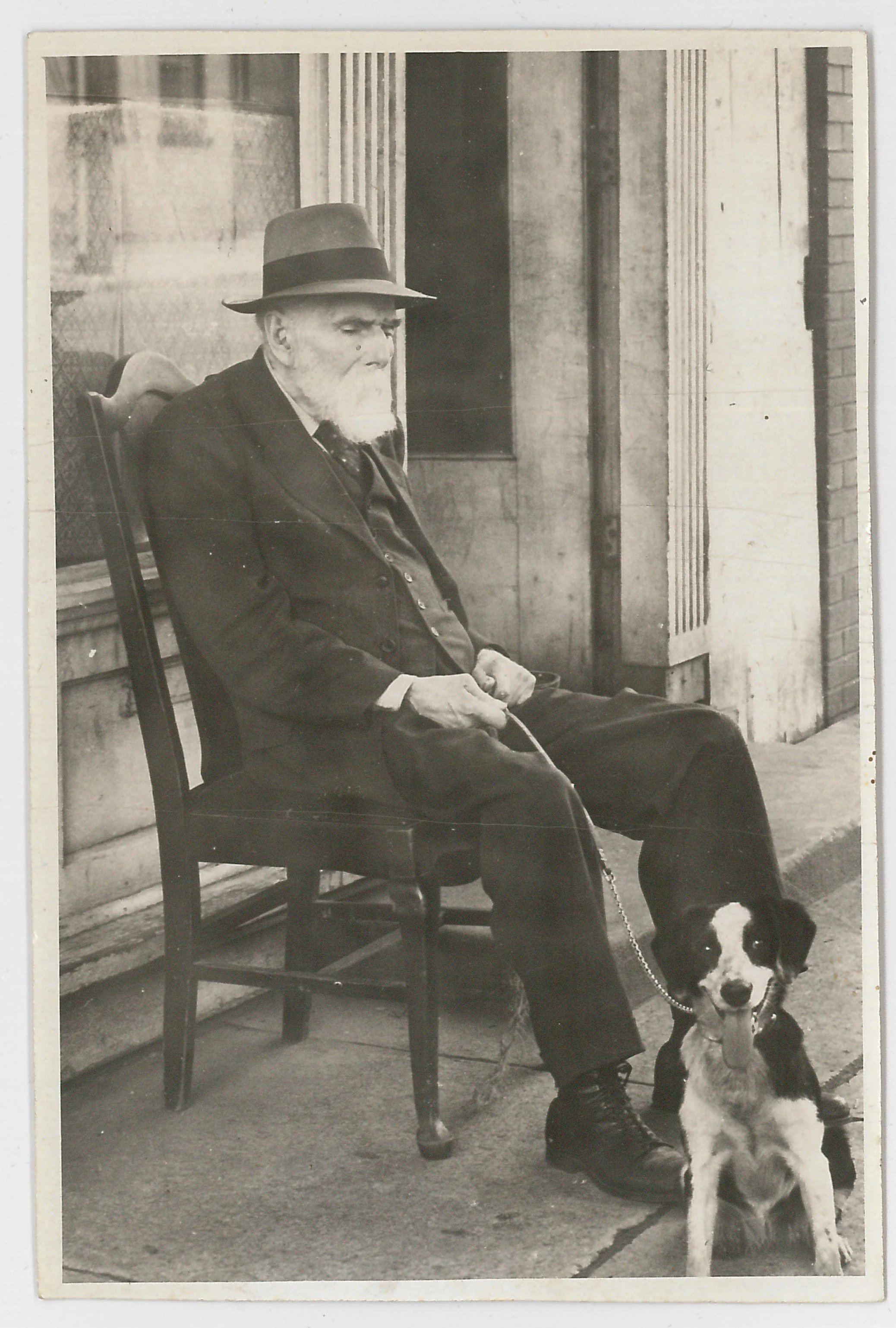 Craig Shaw with a beard and mustache sitting on a wooden chair on a sidewalk, wearing a hat and dark clothing, holding a chain attached to a dog sitting beside him.