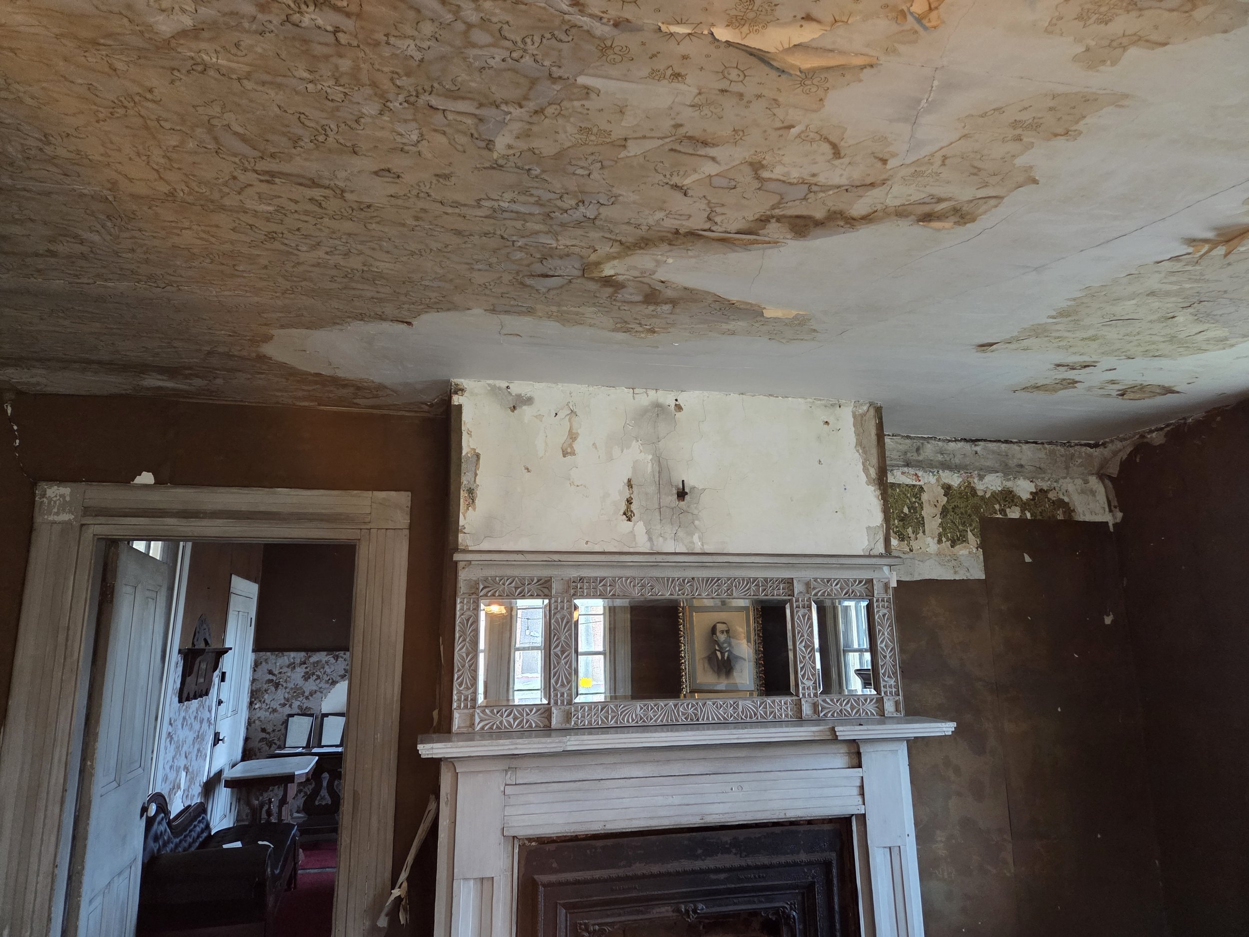 A room with peeling wallpaper  on the ceiling, an ornate white fireplace mantel, and a mirror above it reflecting a portrait painting. There is a doorway leading to another room visible on the left side.
