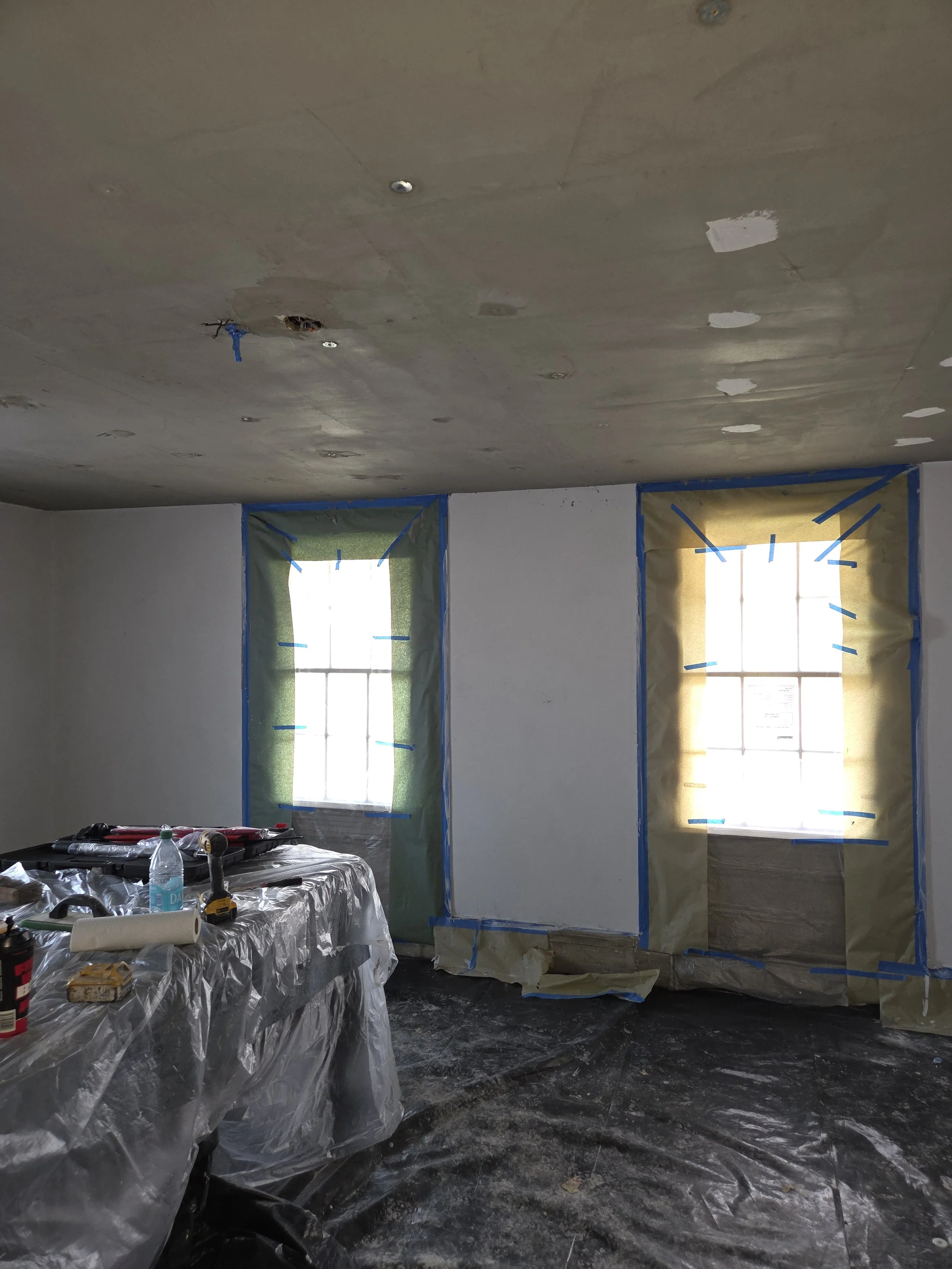 Room under construction with two windows covered with plastic and tape, ceiling partially finished, tools and supplies on a table covered with plastic, floor covered with plastic tarp.