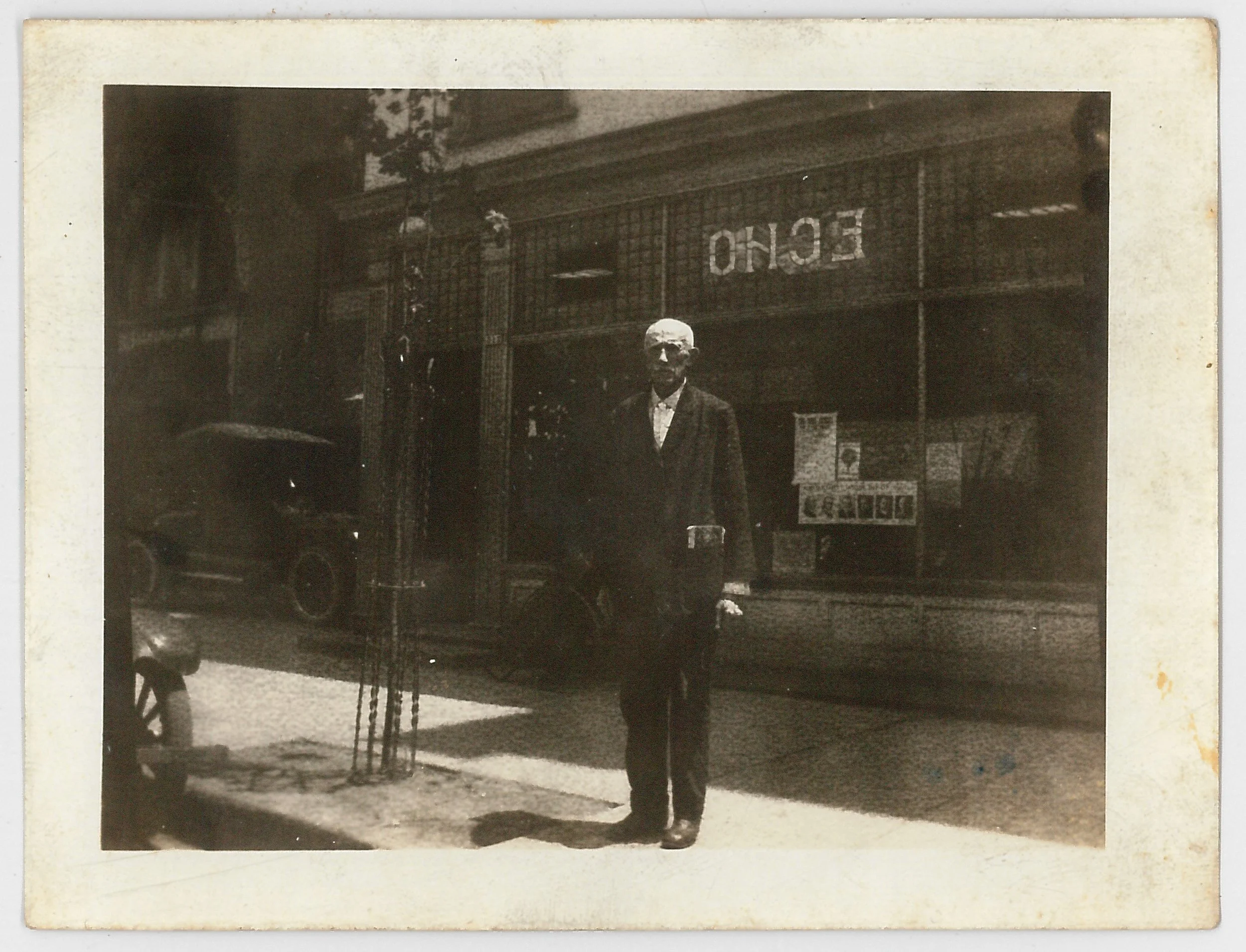 An old black-and-white photograph of a man standing on a sidewalk in front of a shop with a large sign that reads 'ENCHO' in reverse. The man is dressed in a suit and tie, with glasses, and looking toward the camera. A vintage car is partially visible on the left side, and there is a small tree surrounded by a metal grate near the man.