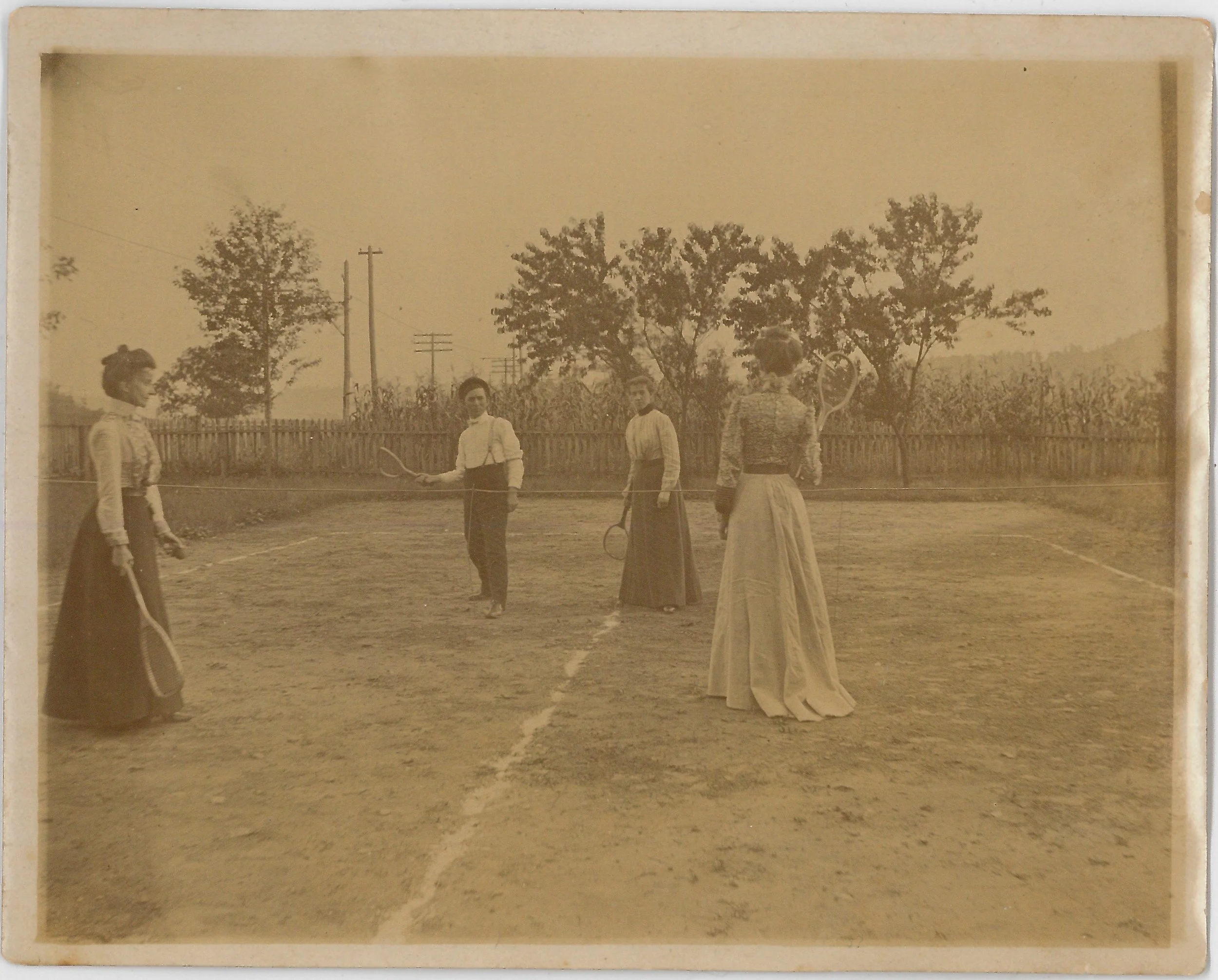 Four women and one man standing on a dirt tennis court, holding tennis rackets, with a wooden fence and trees in the background.