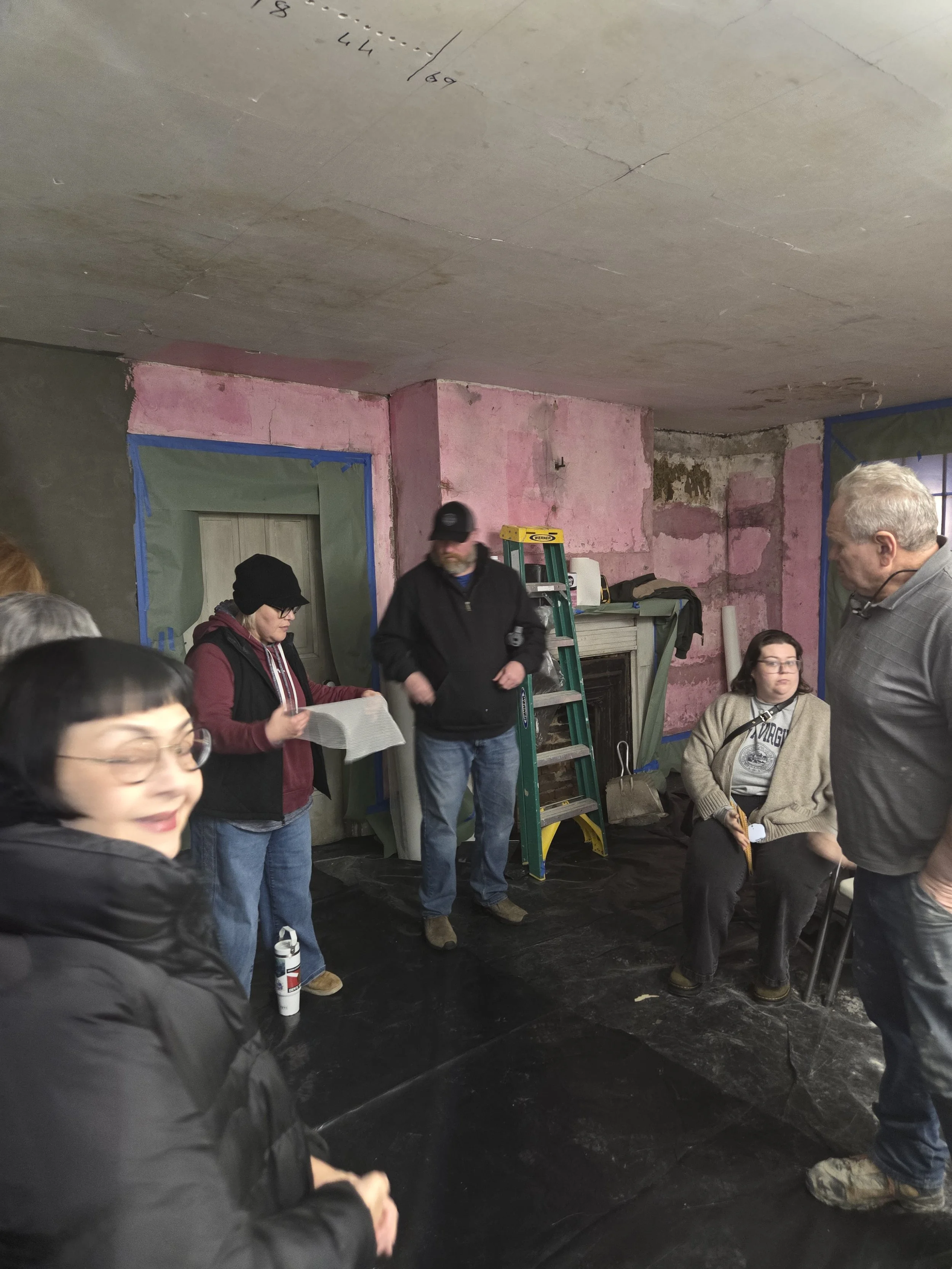 Group of people gathered indoors, some seated and some standing, in a room with unfinished pink and gray walls, a ladder, and supplies.
