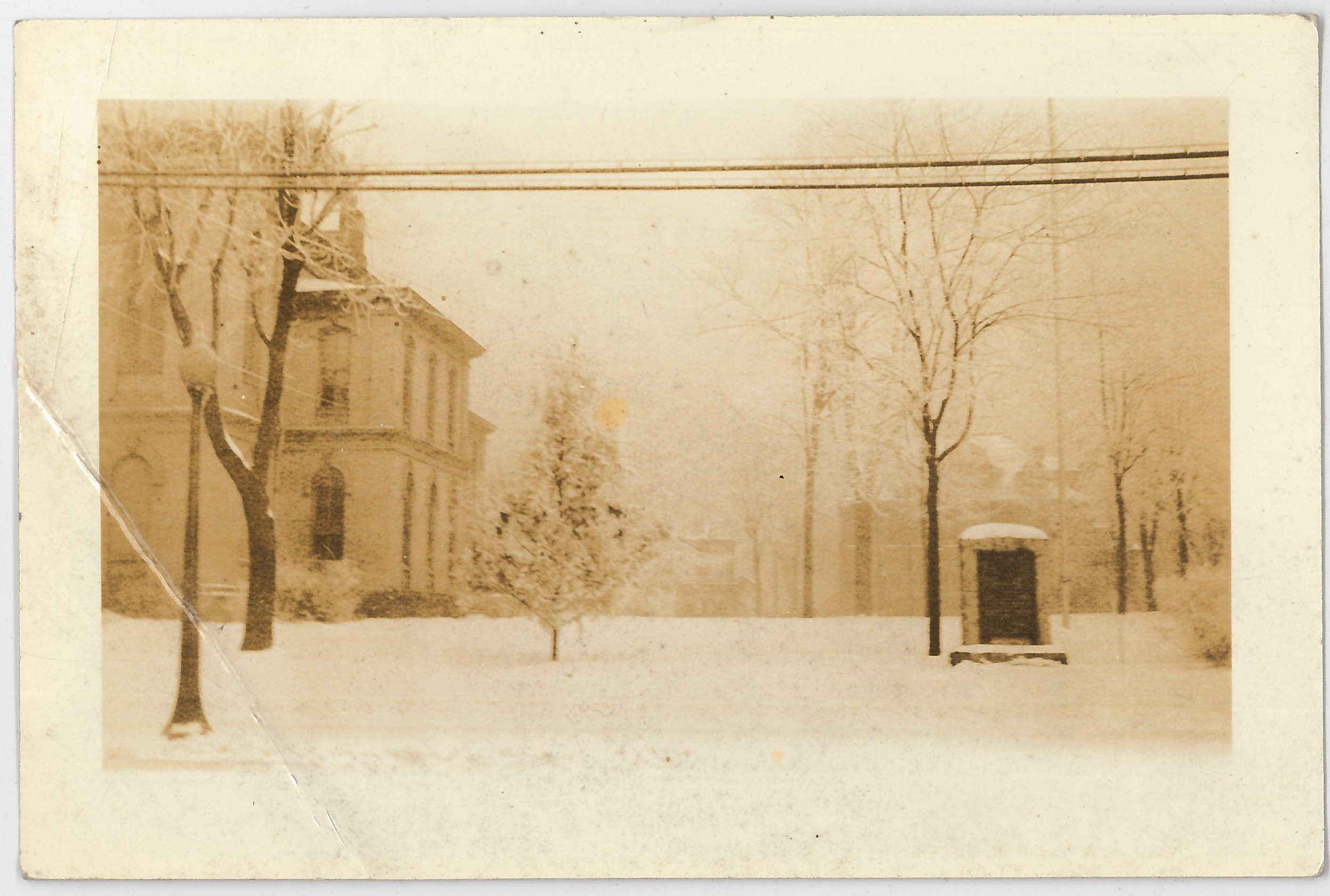 Sepia-toned photograph of a snowy street scene with a large house, leafless trees, and a small shed or kiosk in the background.