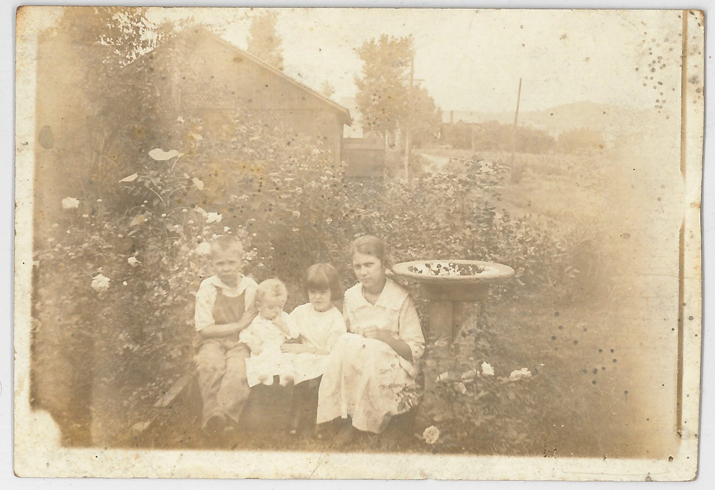 An old sepia-toned photograph of four children sitting on a bench in a garden, with a birdbath to the right and a house with trees and a fence in the background.