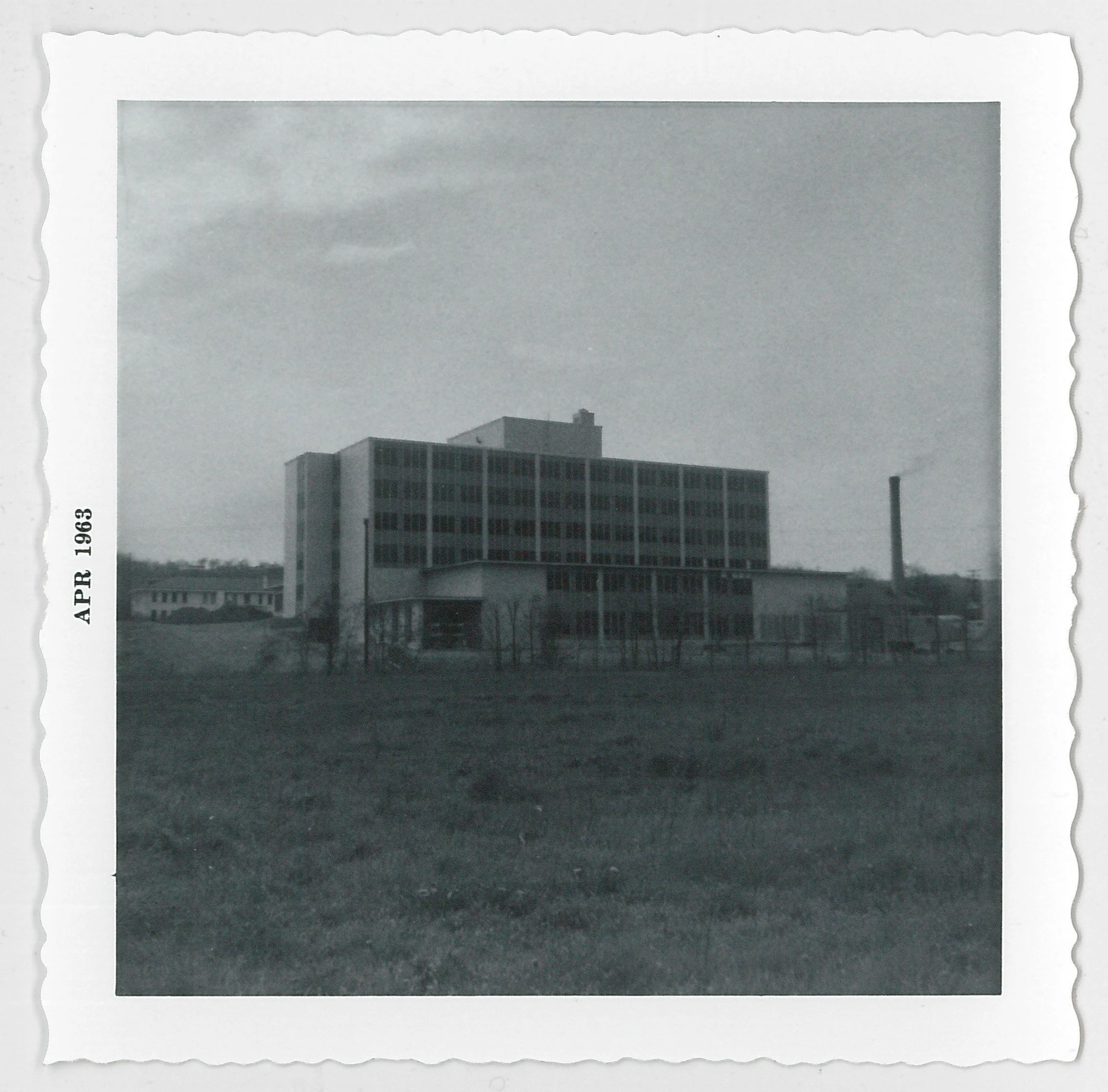 Black and white photograph of a large, multi-story building with a grassy foreground, surrounded by a fence, with a chimney emitting smoke in the background. The photo is dated April 1963.