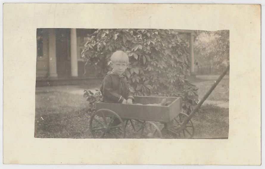 Black and white photograph of a young boy standing next to a toy wagon outdoors, with a bush and a house in the background.