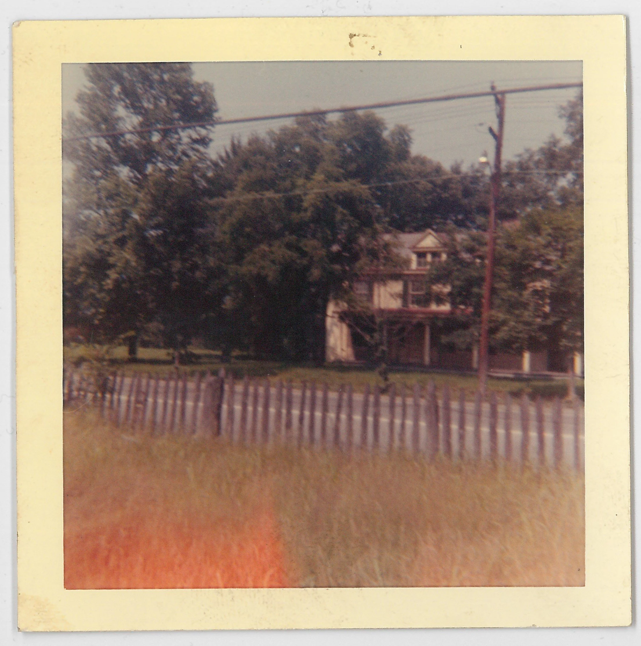 A vintage Polaroid photo of a tree-covered house with a porch, standing behind a wooden fence along a grassy area. Power lines run across the top of the image.