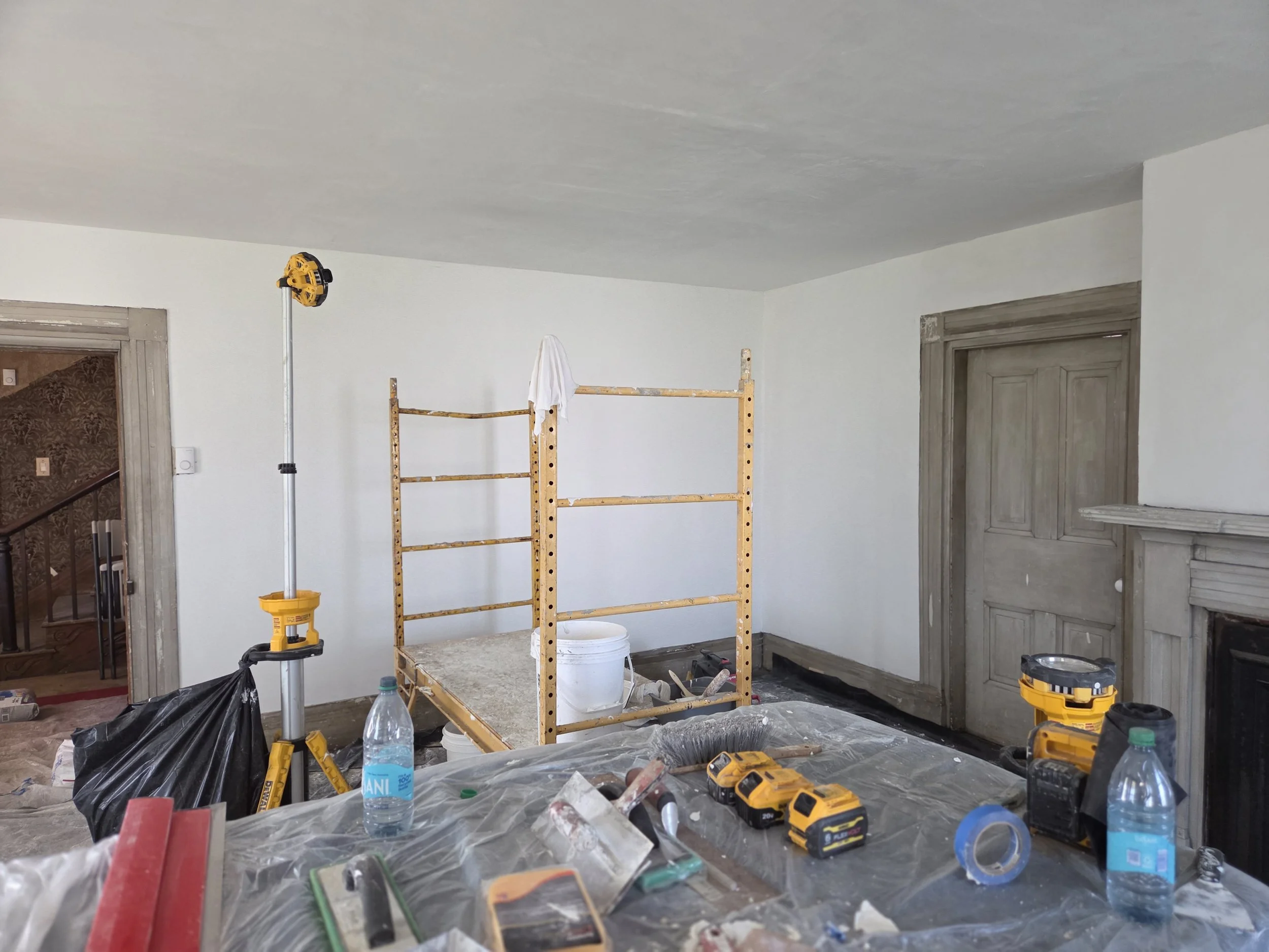 Room undergoing renovation with tools, water bottles, and construction equipment scattered on a plastic-covered surface, a wooden scaffold in the center, and an unfinished wall and doorway visible.