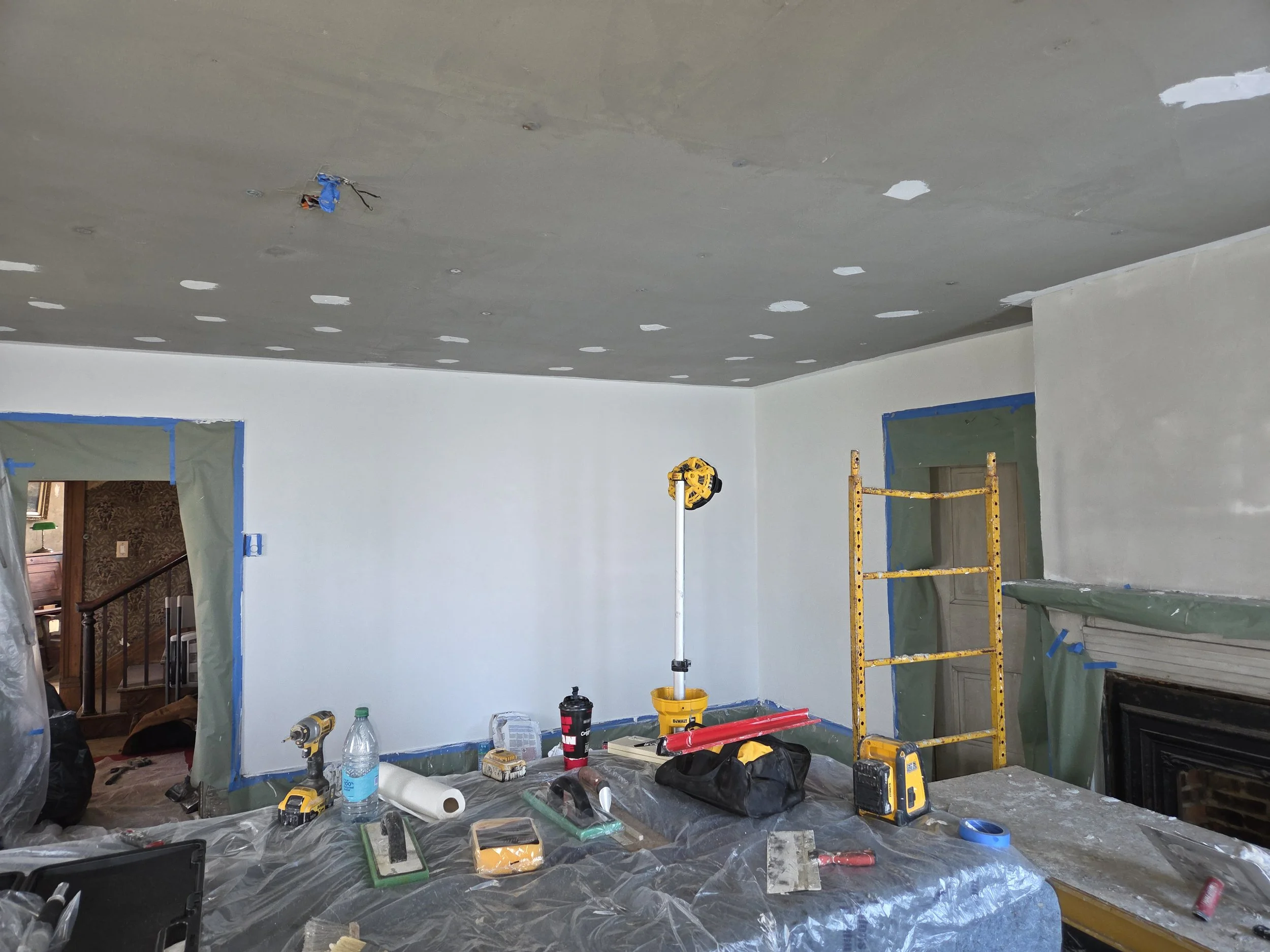 Room under renovation with tools and equipment, including ladder, drill, tape measure, paint supplies scattered on a table, and new drywall installed.