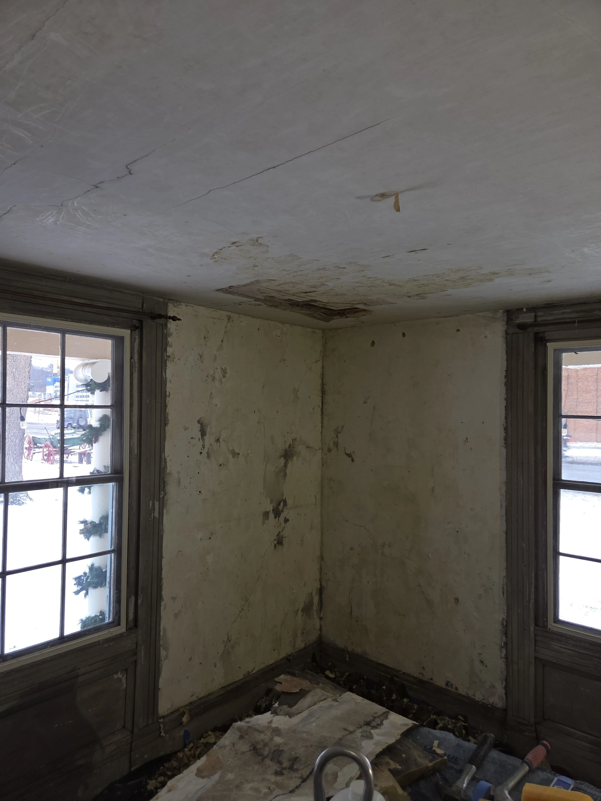Empty, dilapidated room with peeling wallpaper, cracked walls, and a damaged ceiling. Two windows with trim are visible, showing a snowy outdoor scene with some decorations on the window panes.