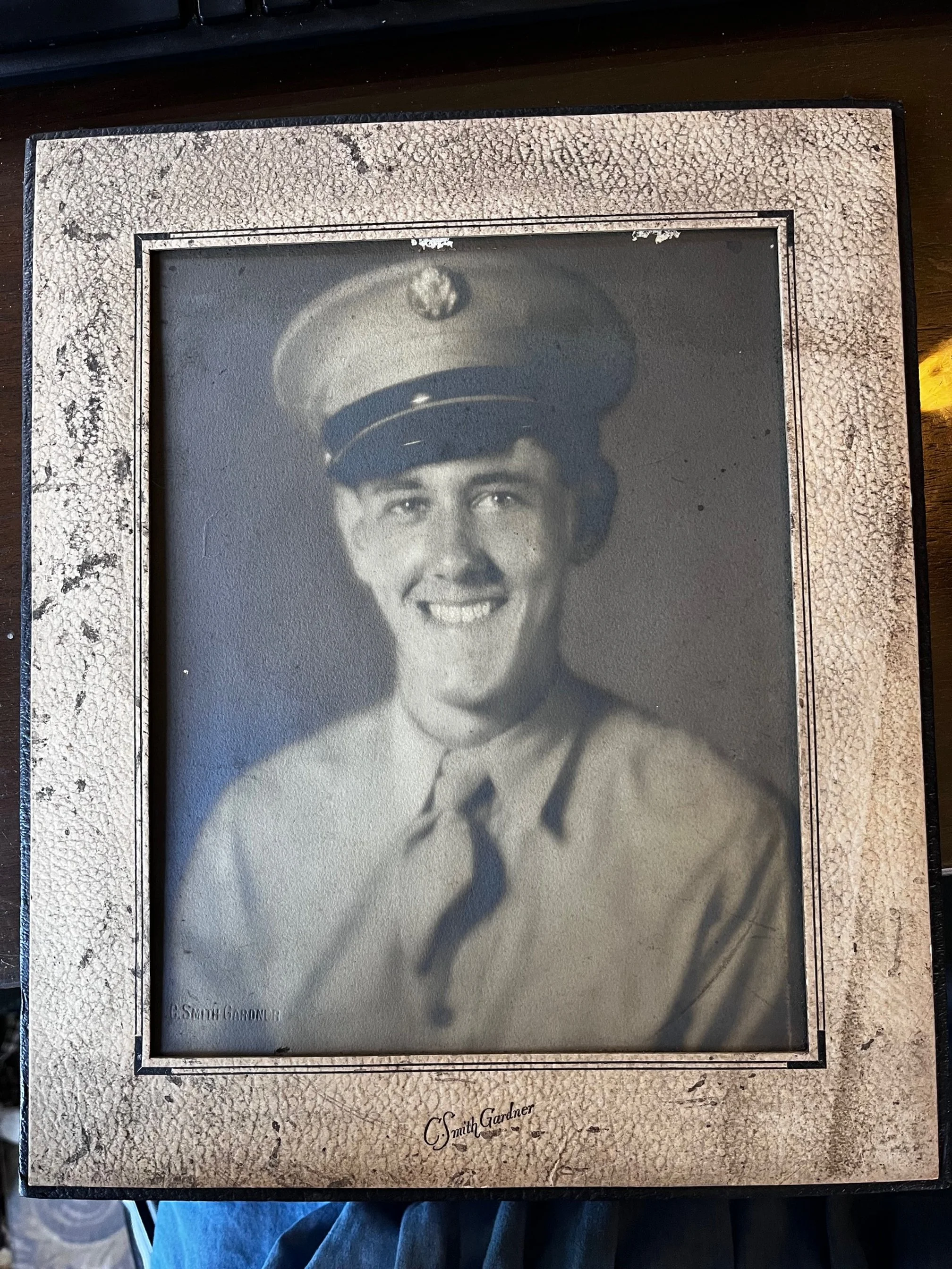 Black-and-white portrait of a smiling young man in military uniform and cap, framed in a weathered display case.
