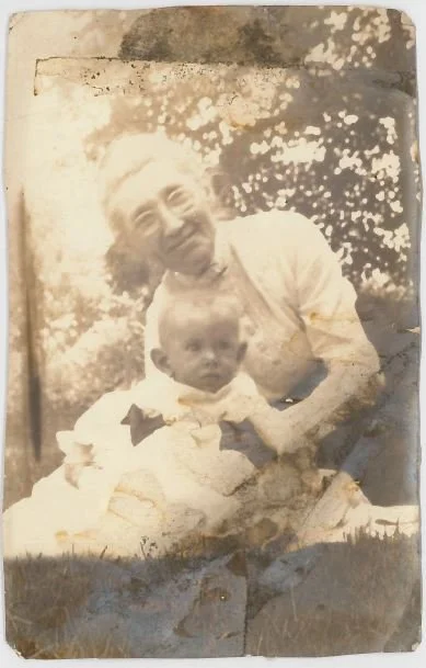 A sepia-toned photo of an elderly woman with glasses holding a young child outdoors, with trees in the background.