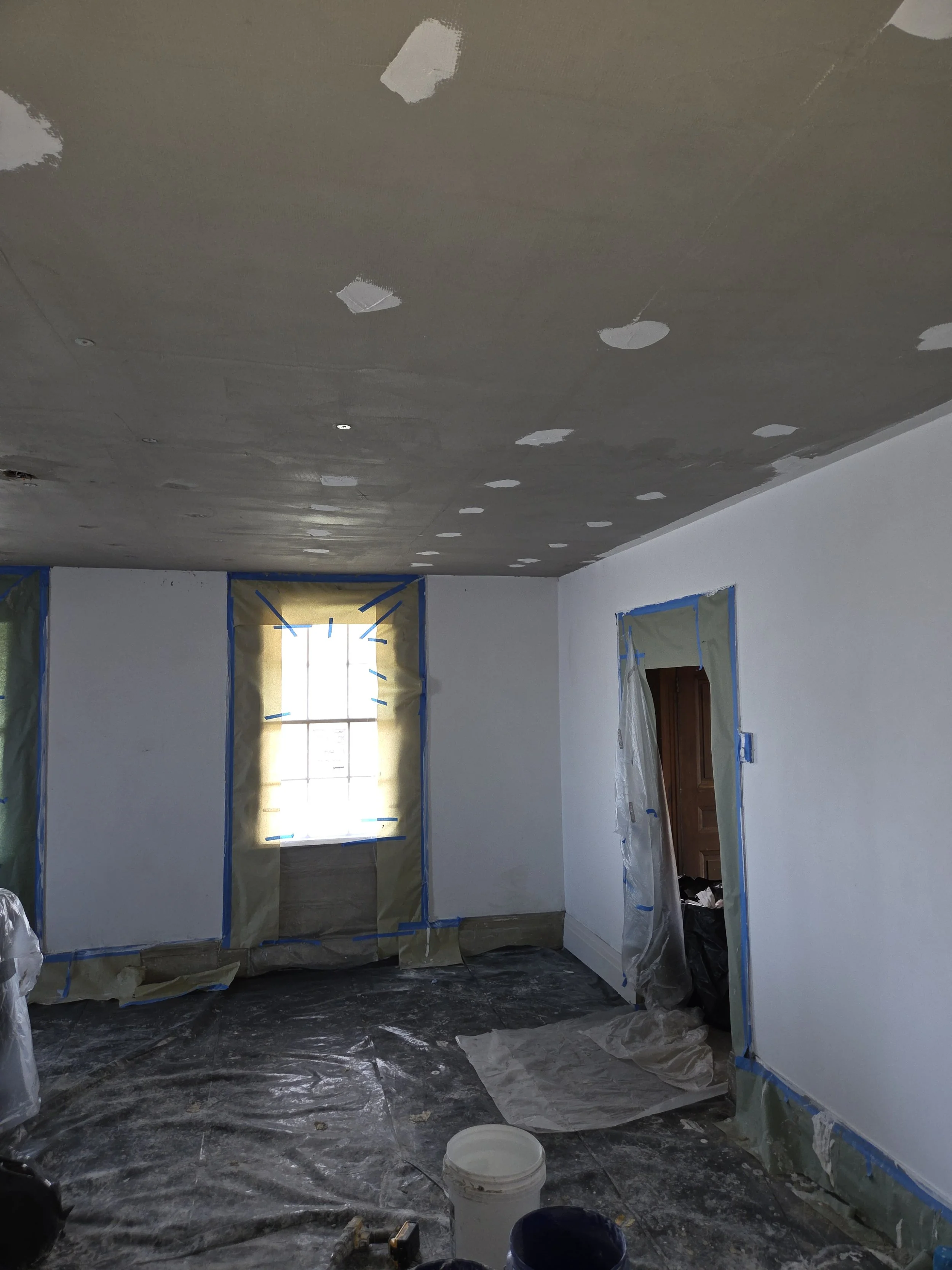 A room under renovation with a window and doorway covered in painter's plastic, a ceiling with location marks, and a floor covered in plastic sheeting and tools.