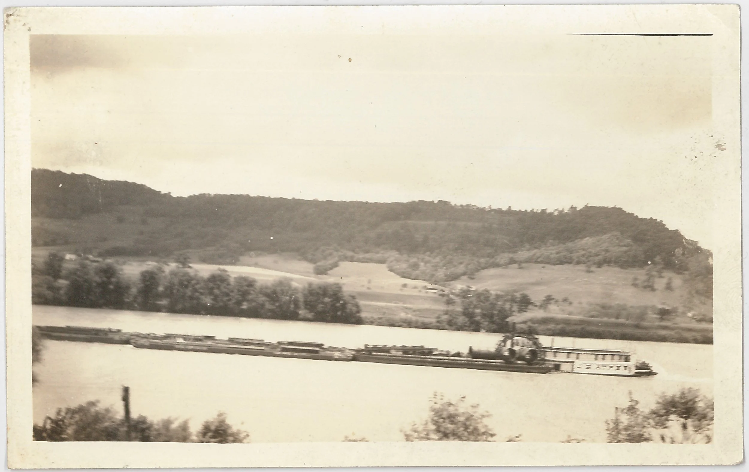 Old black and white photograph of a river with a barge carrying a tractor and a flatbed trailer moving on calm water, with rolling hills and trees in the background.