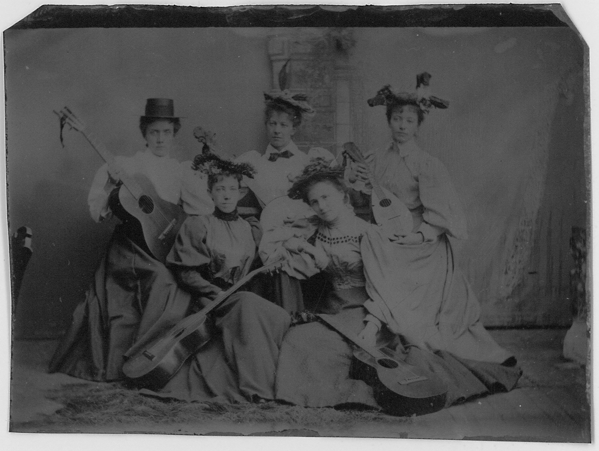 A black and white vintage photo of six women in historical costumes, some holding guitars and a long sword, posed for a group portrait.