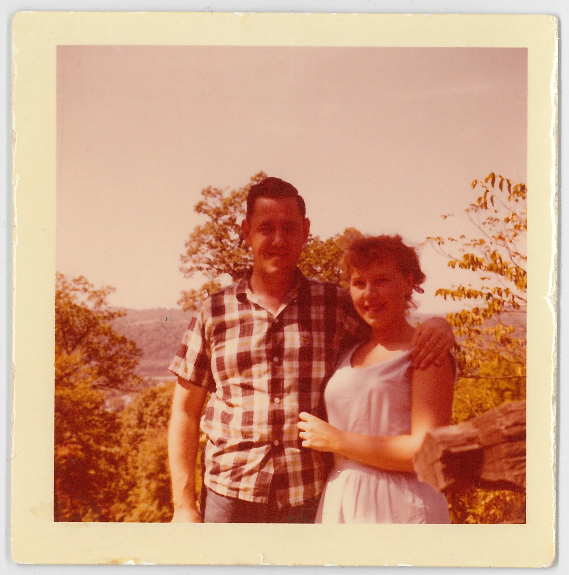 A vintage color photograph of John and Lisa Cockayne standing outdoors by a wooden fence during daytime, with trees and hills in the background, smiling at the camera.