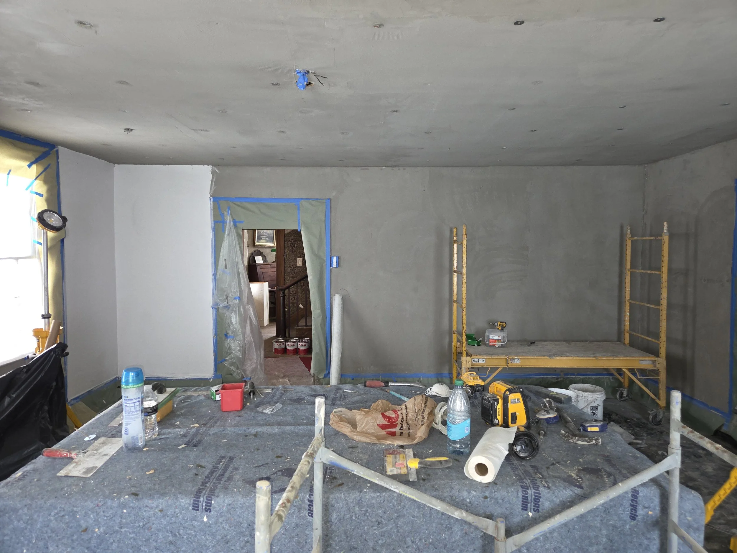 Room under renovation with plaster being applied, tools, and construction materials on a table, with an opening to another room in the background.