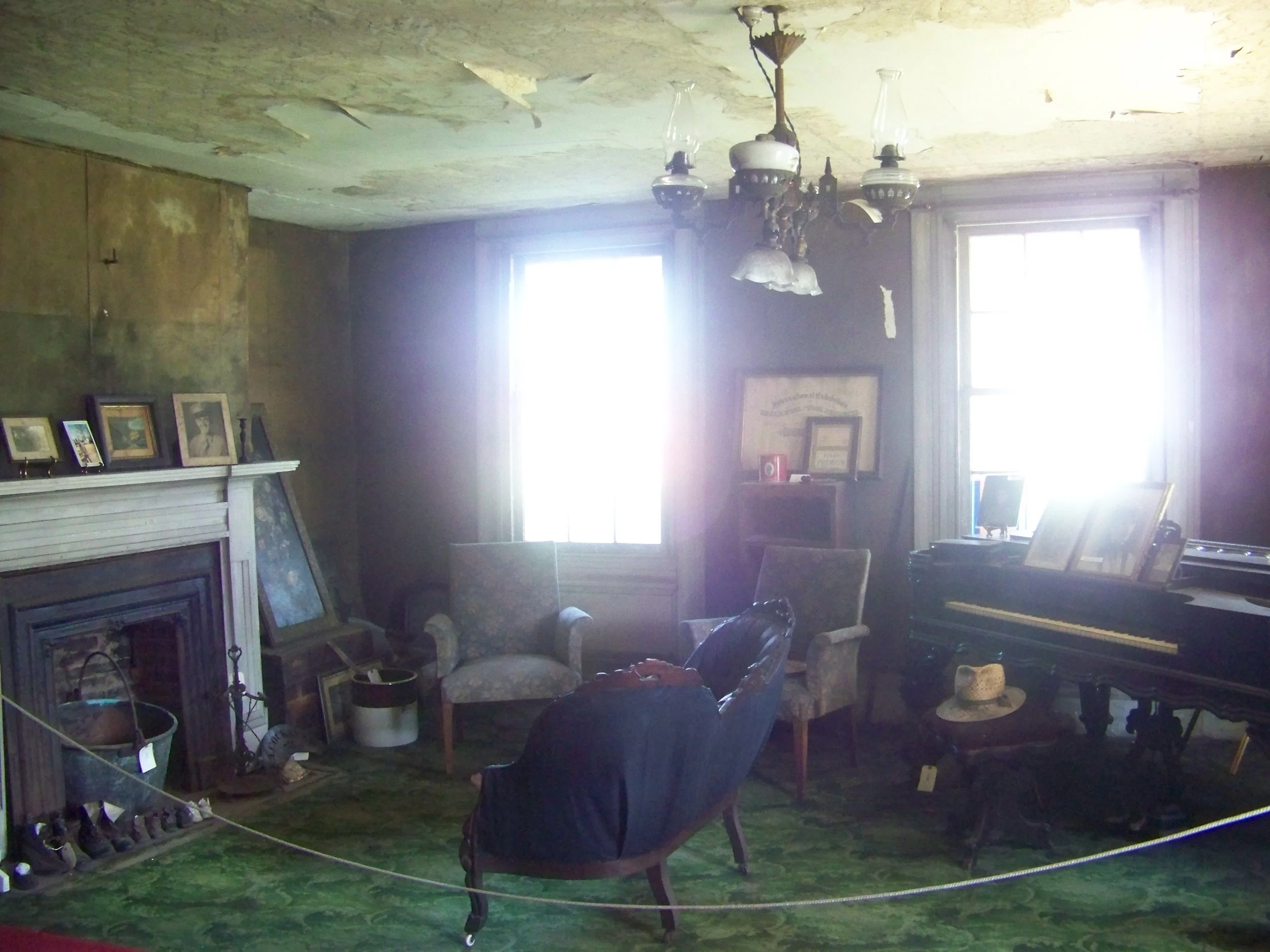 A vintage living room with an old chandelier, worn ceiling, and peeling walls. The room contains a black piano with framed photos, a straw hat on a table, and old-fashioned armchairs. Sunlight streams through two large windows, illuminating the dusty, cluttered space.