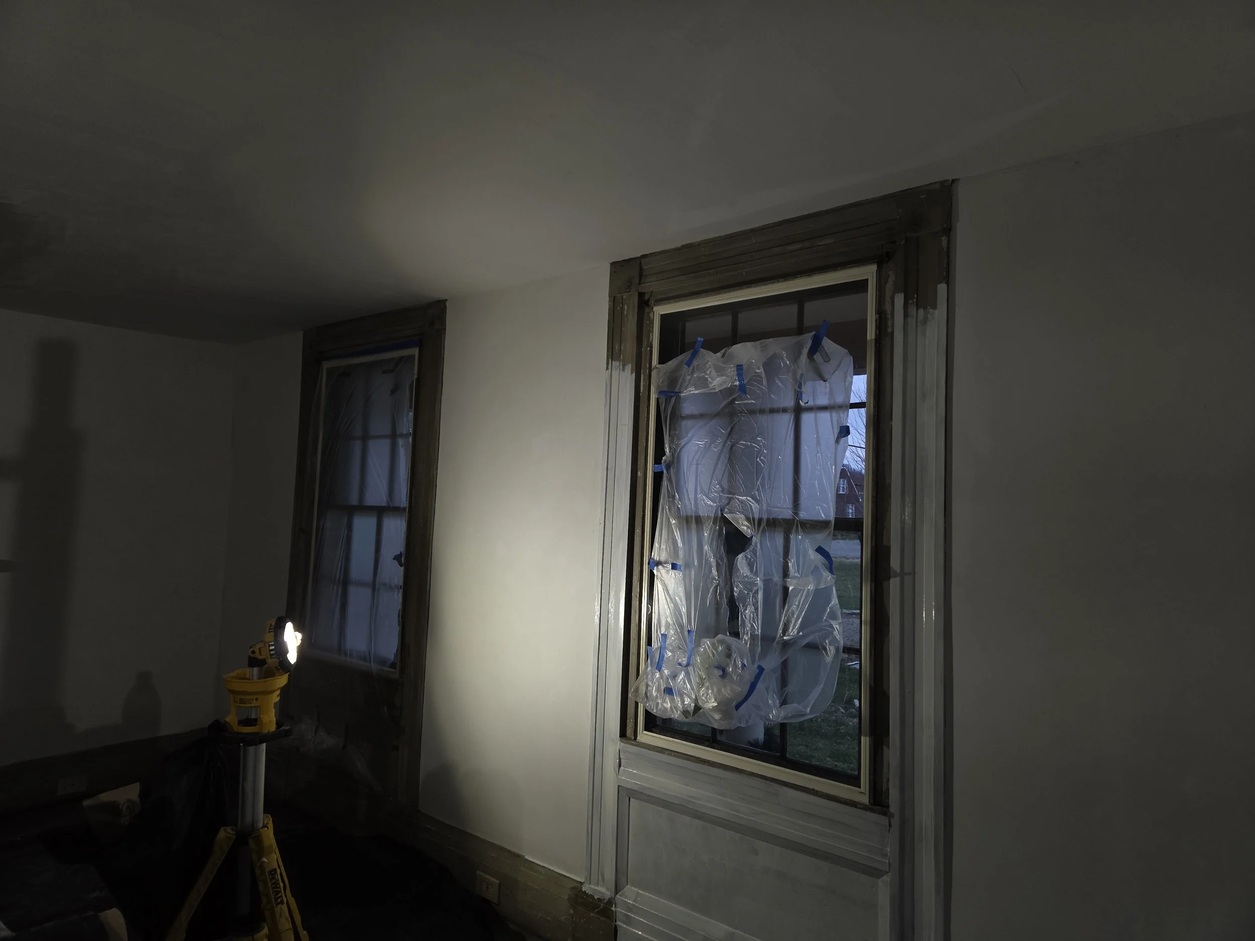 Interior of a room under construction or renovation, with two windows, one of which is covered with plastic fastened by blue painter's tape. A yellow work light is on, illuminating part of the room.