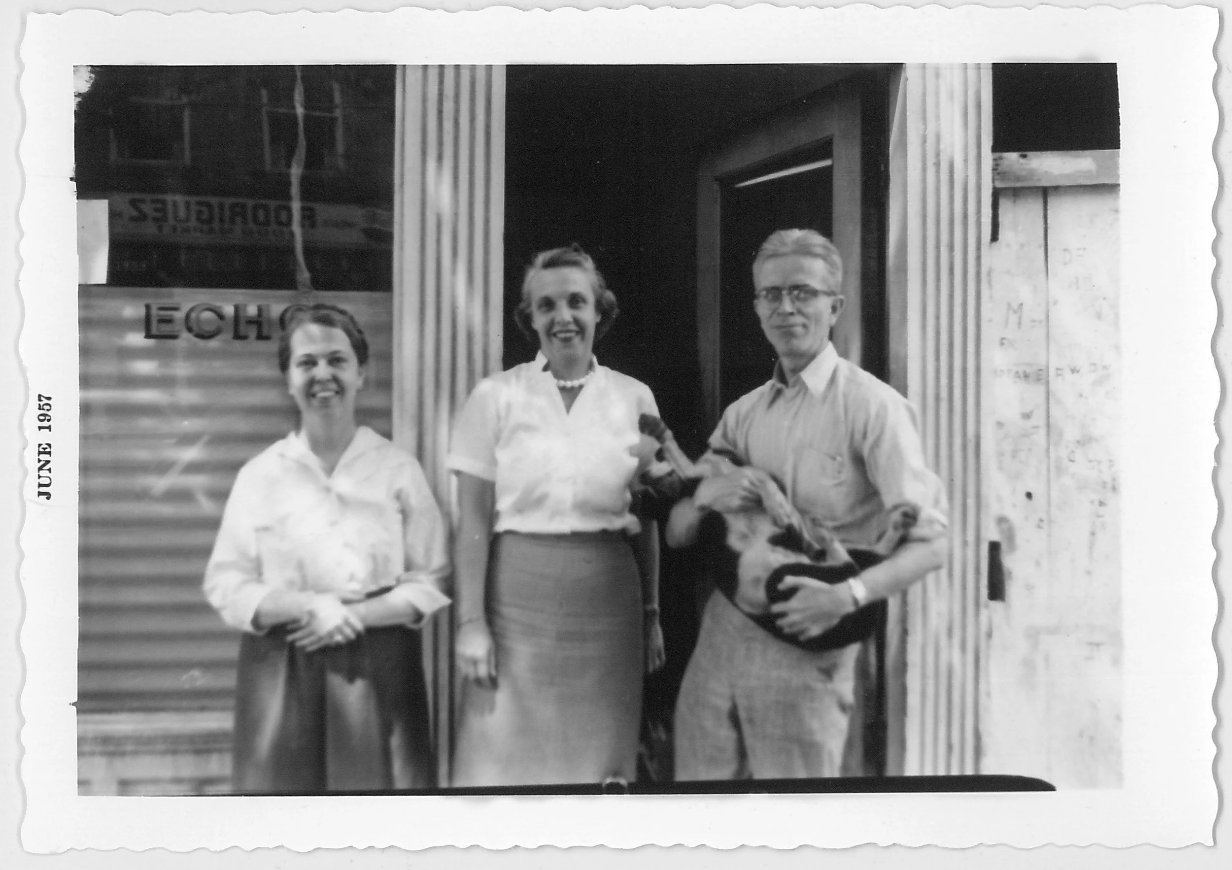 Two women and Sam Shaw standing outside the ECHO in June 1957. The man is holding a dog. The women are smiling, and the women are dressed in skirts and blouses, while the man is in a short-sleeve shirt and trousers.