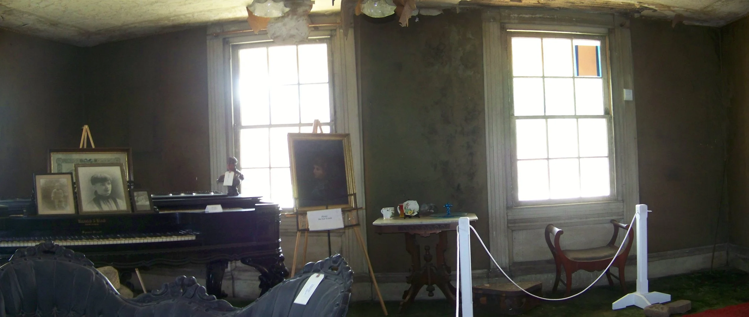 Room with old furniture, framed photographs on a piano, a portrait of a young boy, and a small table with decorative items, in front of large windows with sunlight streaming in.