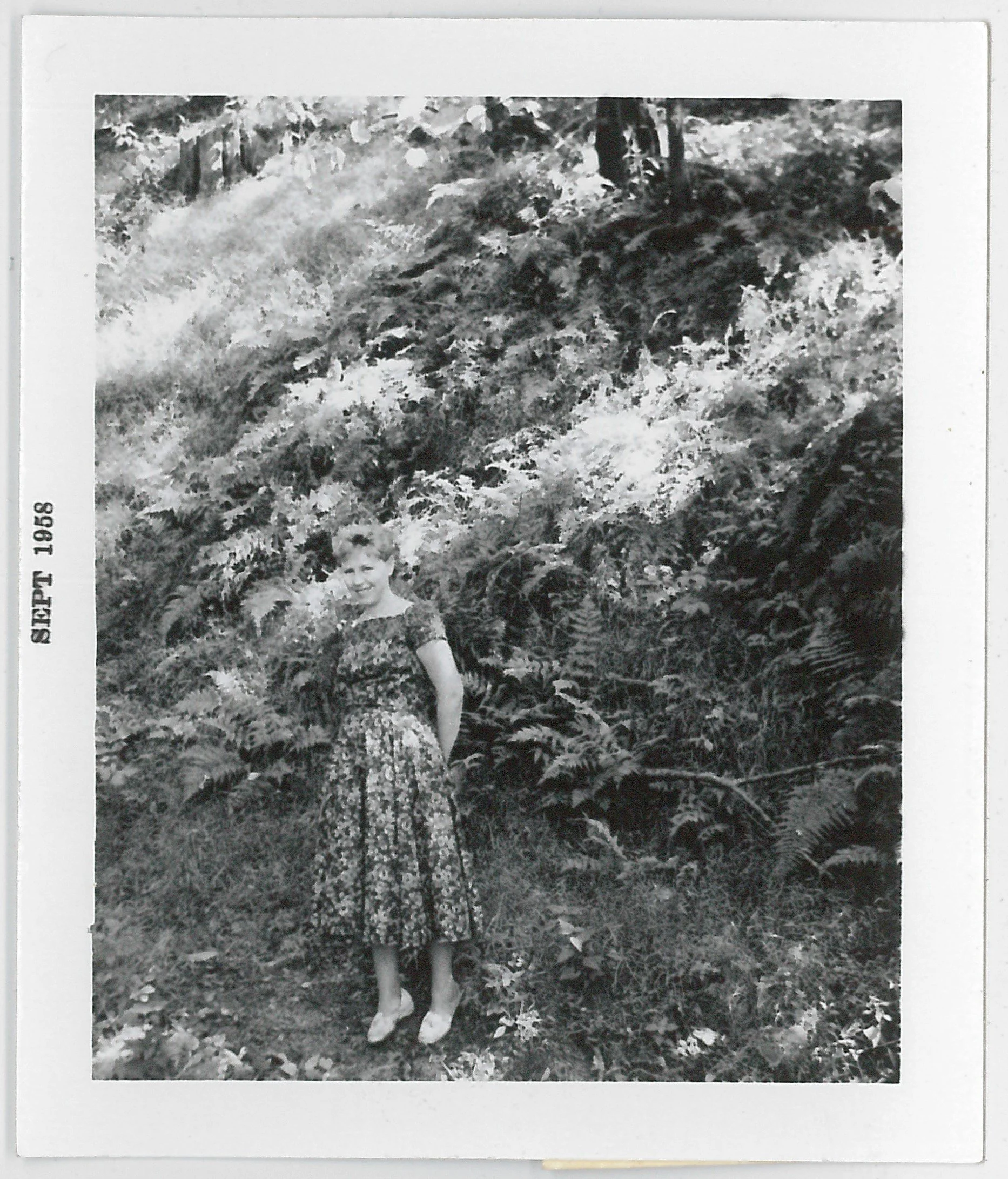A young Lisa Cockayne standing outdoors on grass, wearing a patterned dress and shoes, with ferns and foliage in the background. The date 'September 1958' is printed on the left side.