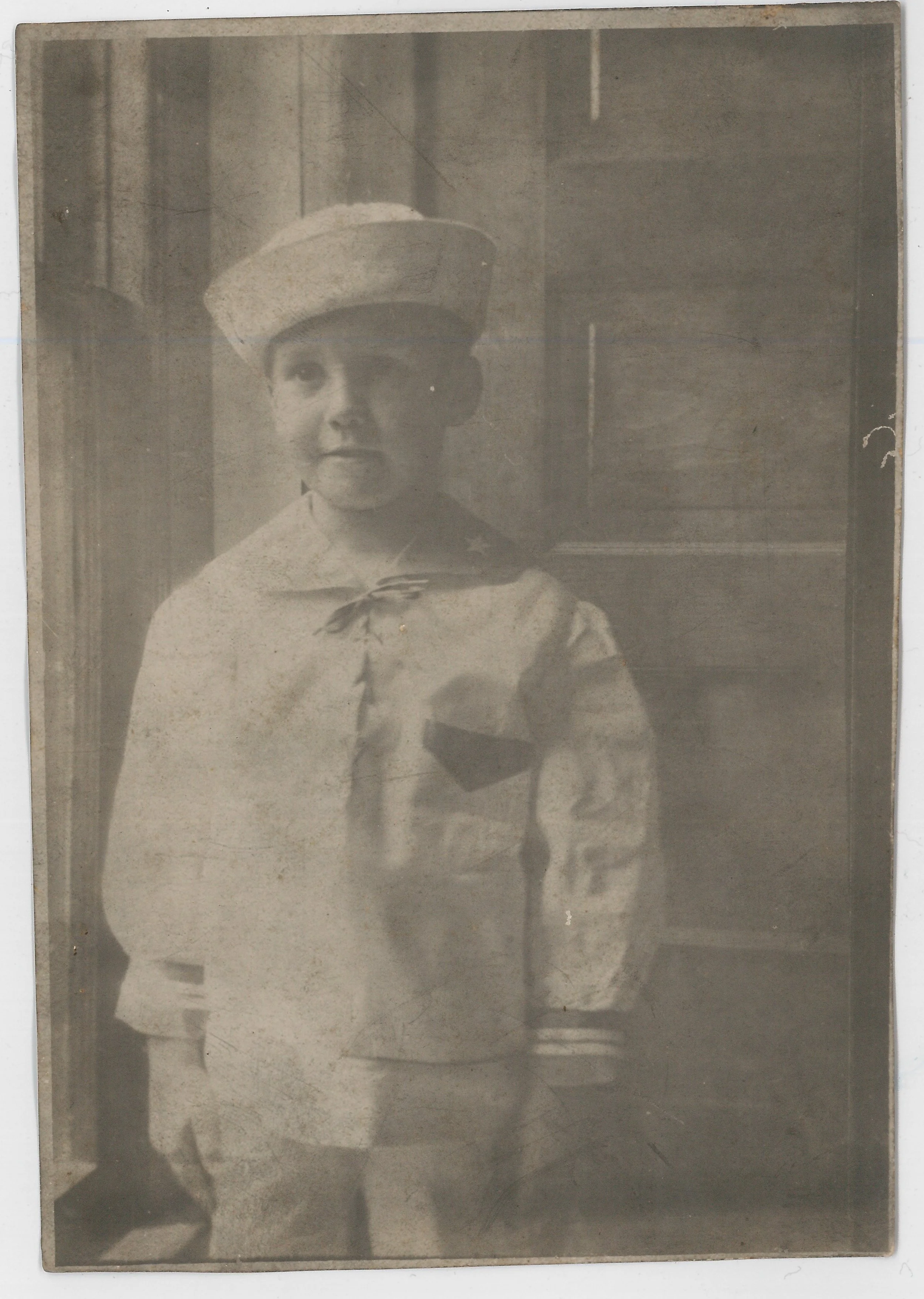 A young John in a scout uniform standing indoors, wearing a hat and a neckerchief with a wooden wall in the background.