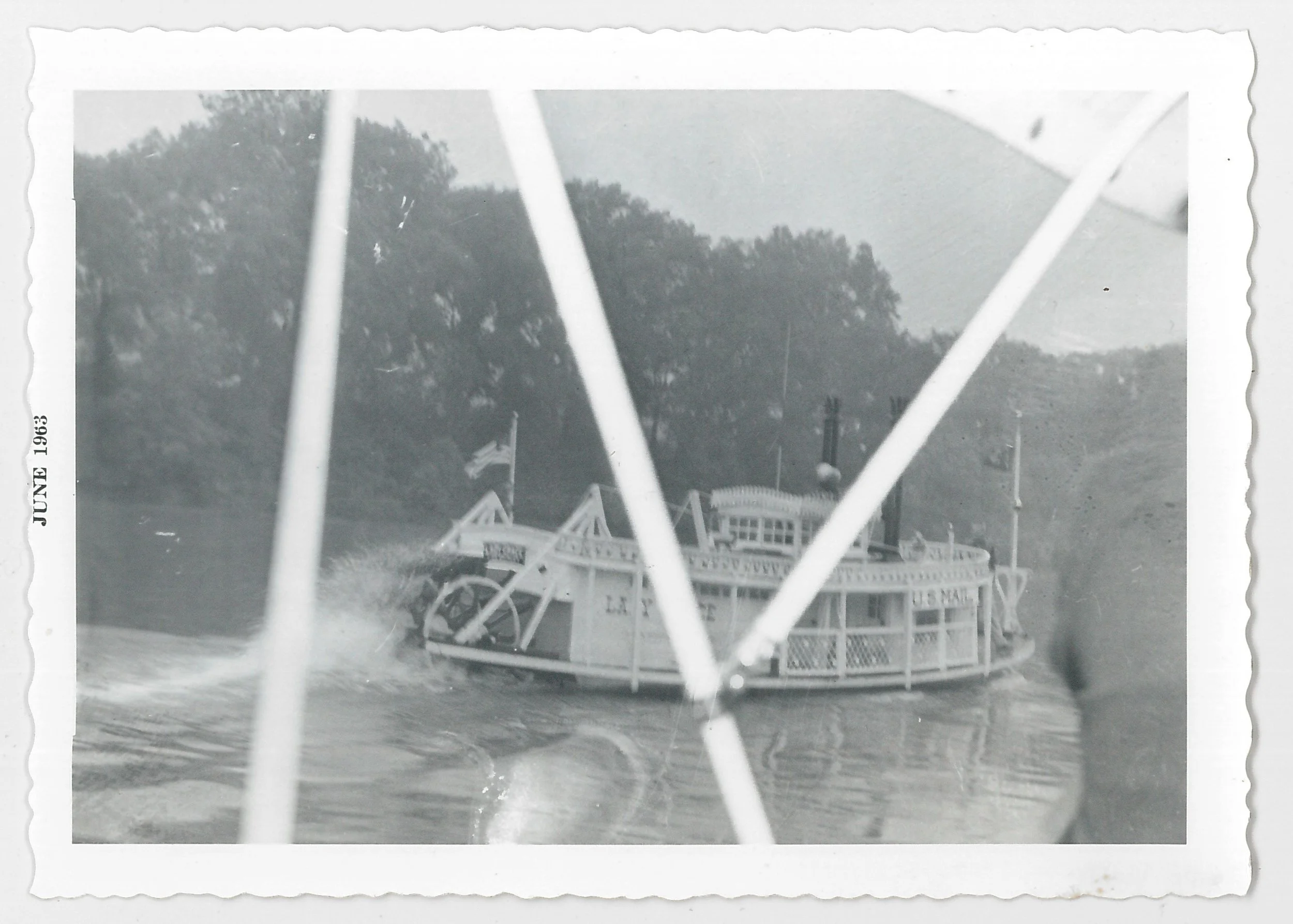 Black-and-white photo of a paddleboat on a river, seen through the boat's railing. Trees line the riverbank in the background. The date