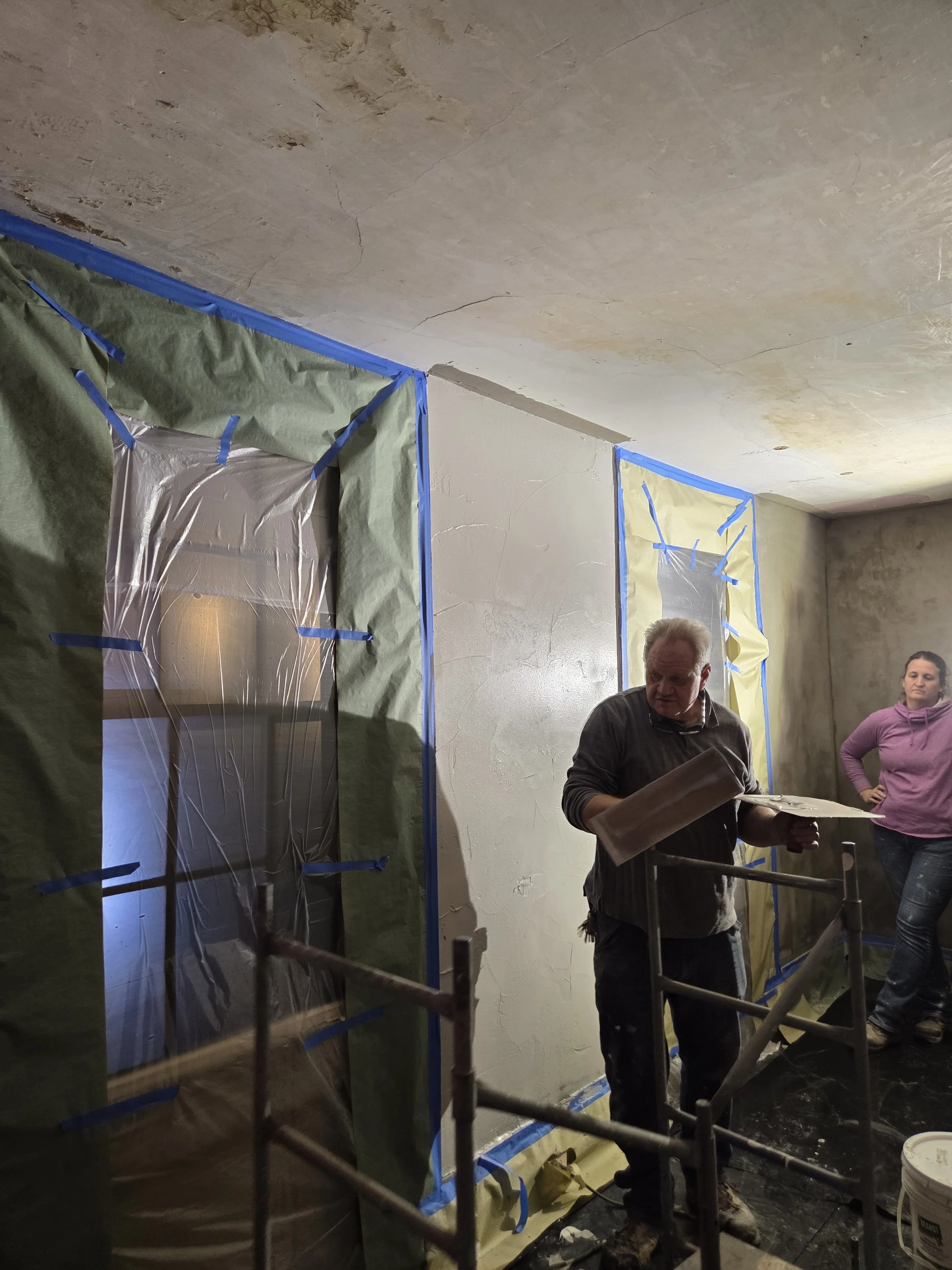 People working on interior wall renovation, with one person holding a plastering tool and standing near plastic-covered window areas taped off for painting or drywall work.