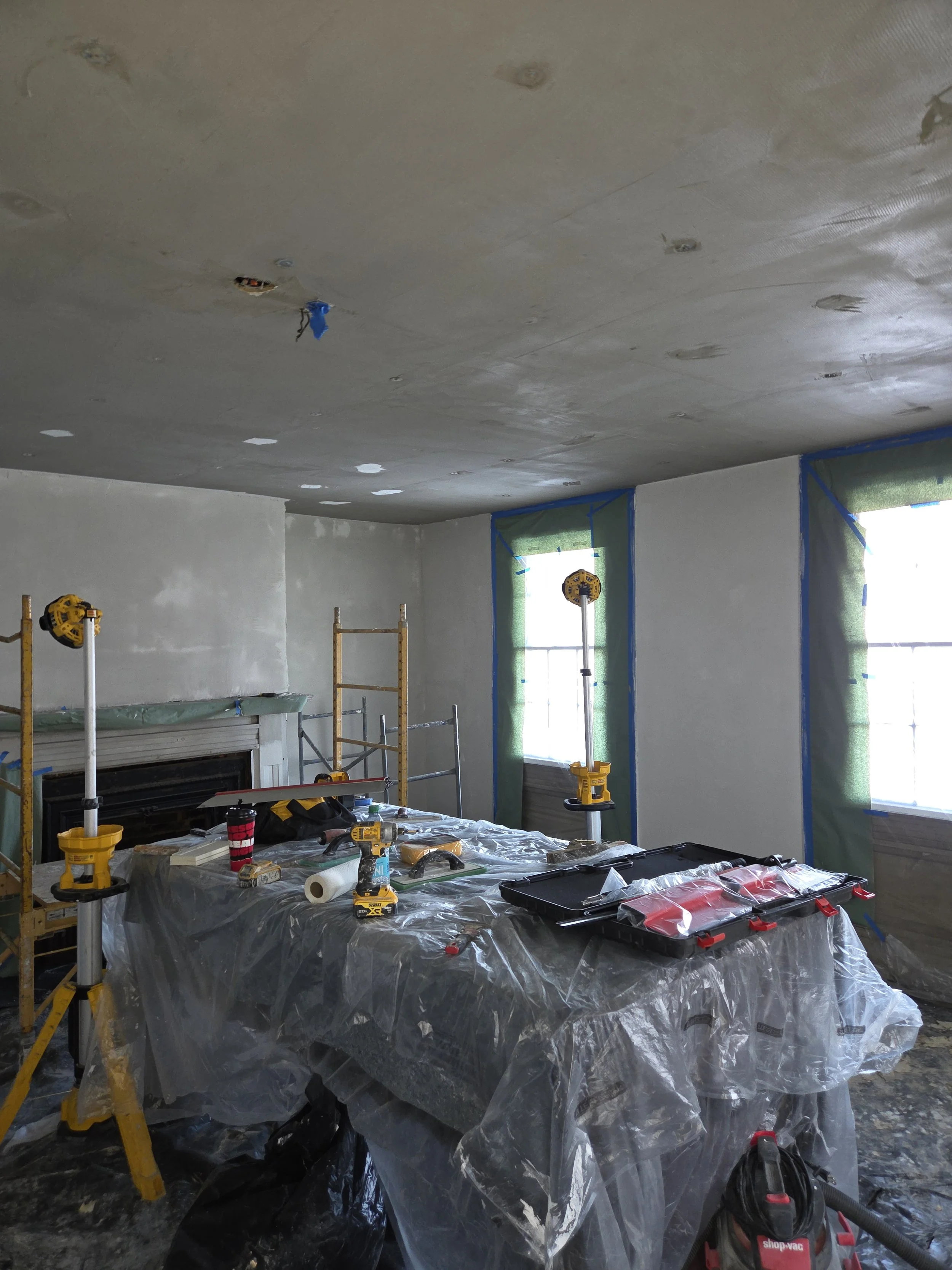 Room under construction with windows covered by green plastic, scaffolding, and a table covered with plastic, construction tools, and equipment.