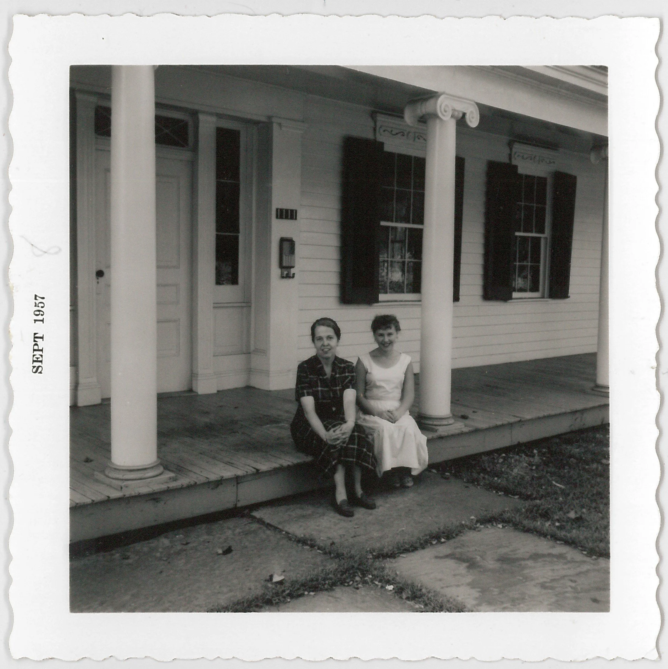 Two women sitting on the porch of a house, smiling, with a white door and windows with shutters behind them. The photo is in black and white, dated September 1957.
