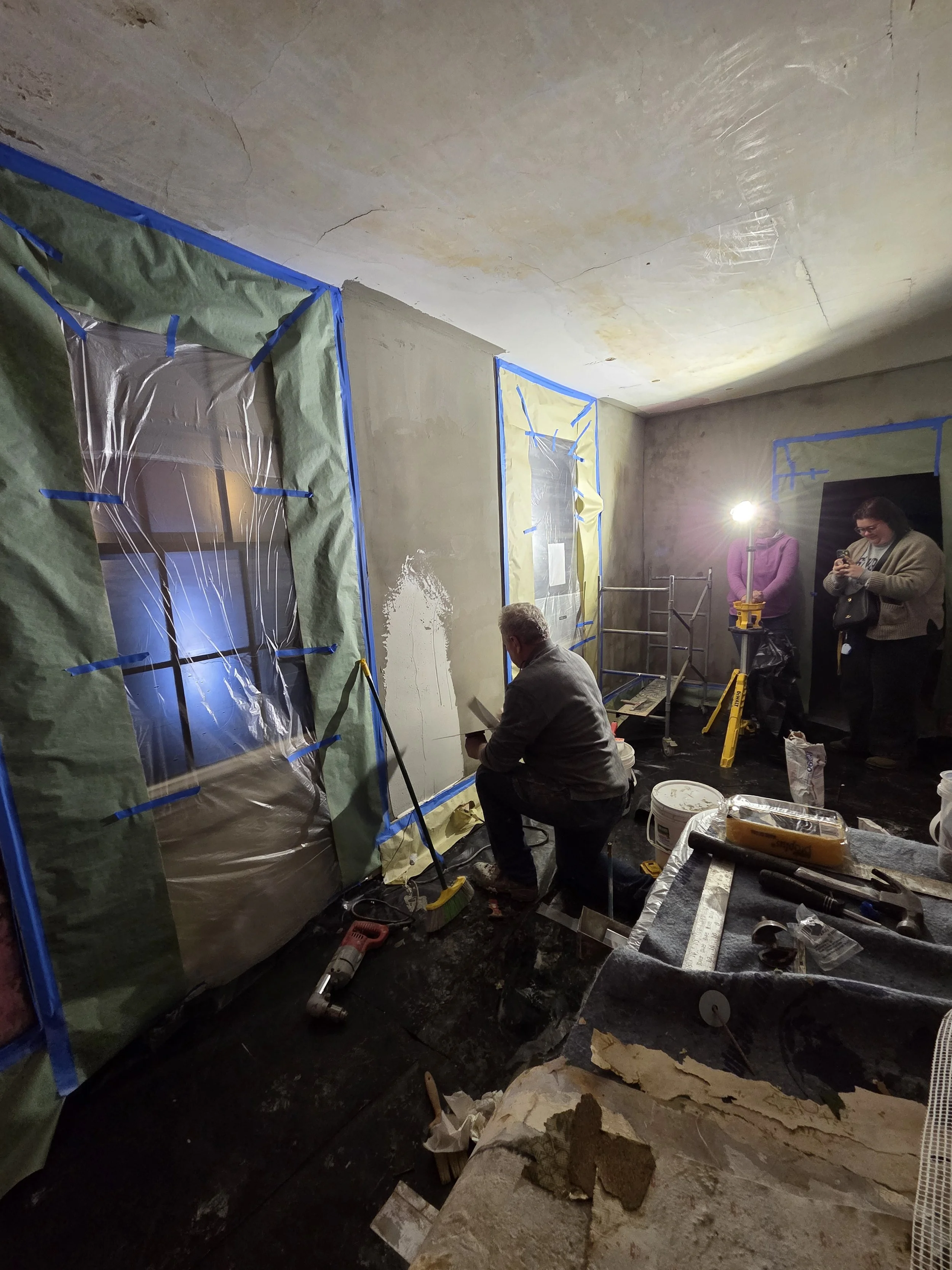 People working on renovation or construction inside a room with taped windows and wall patches, tools, and equipment around.