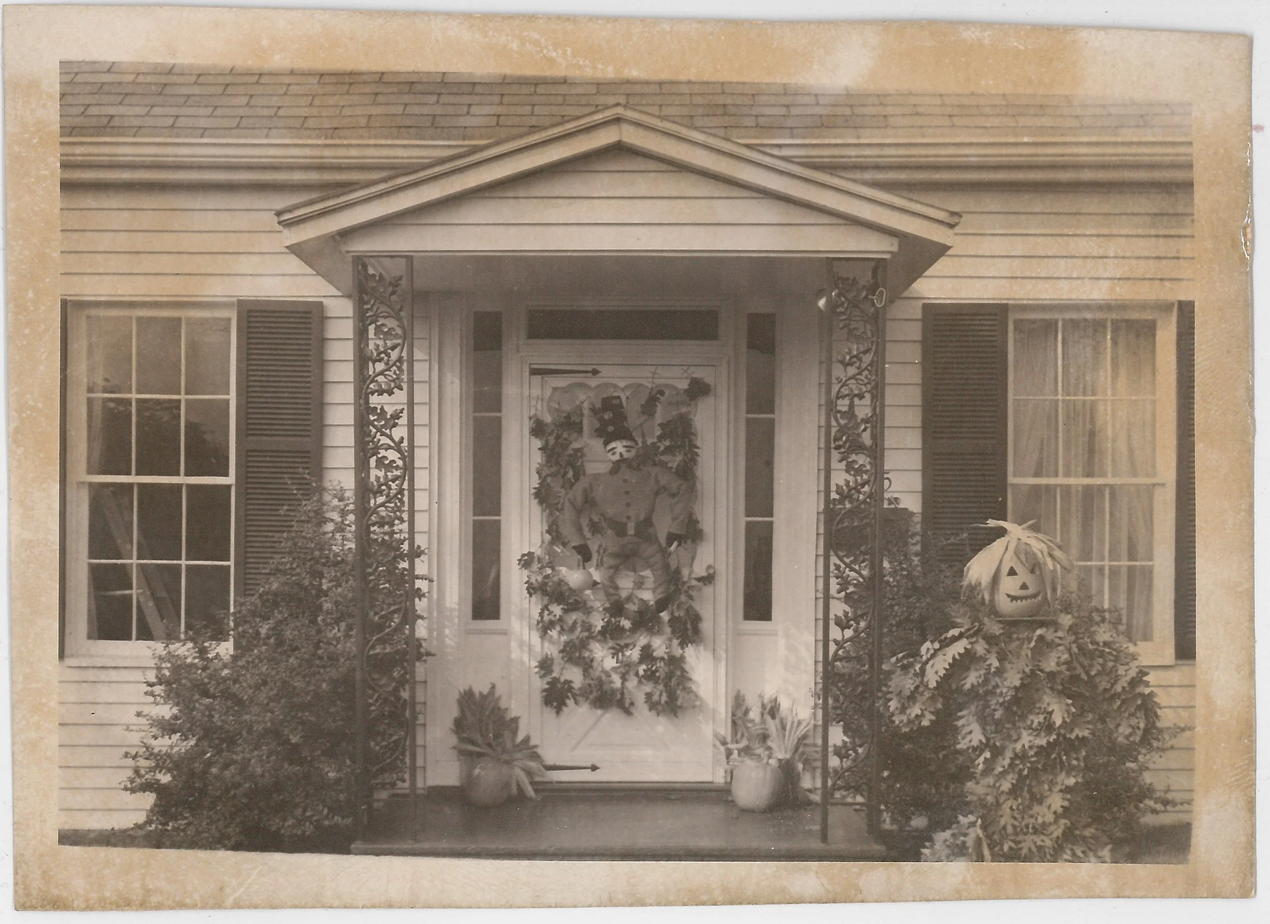 A sepia-toned photo of a house's front porch with a decorated Halloween door featuring a clown, pumpkins, and plants.
