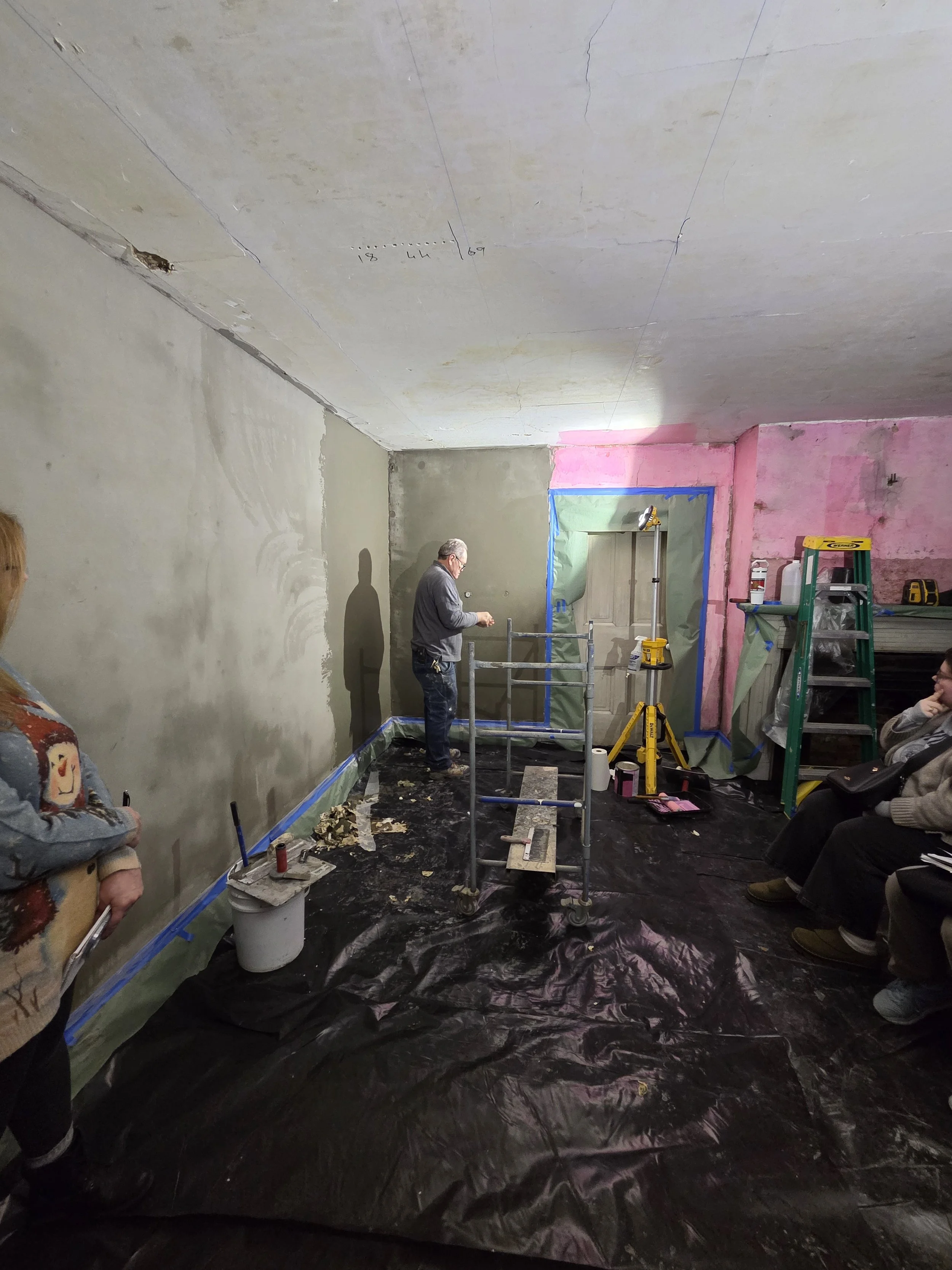 Room under renovation with a worker plastering a wall gray, surrounded by painting supplies and construction tools and ladders around.