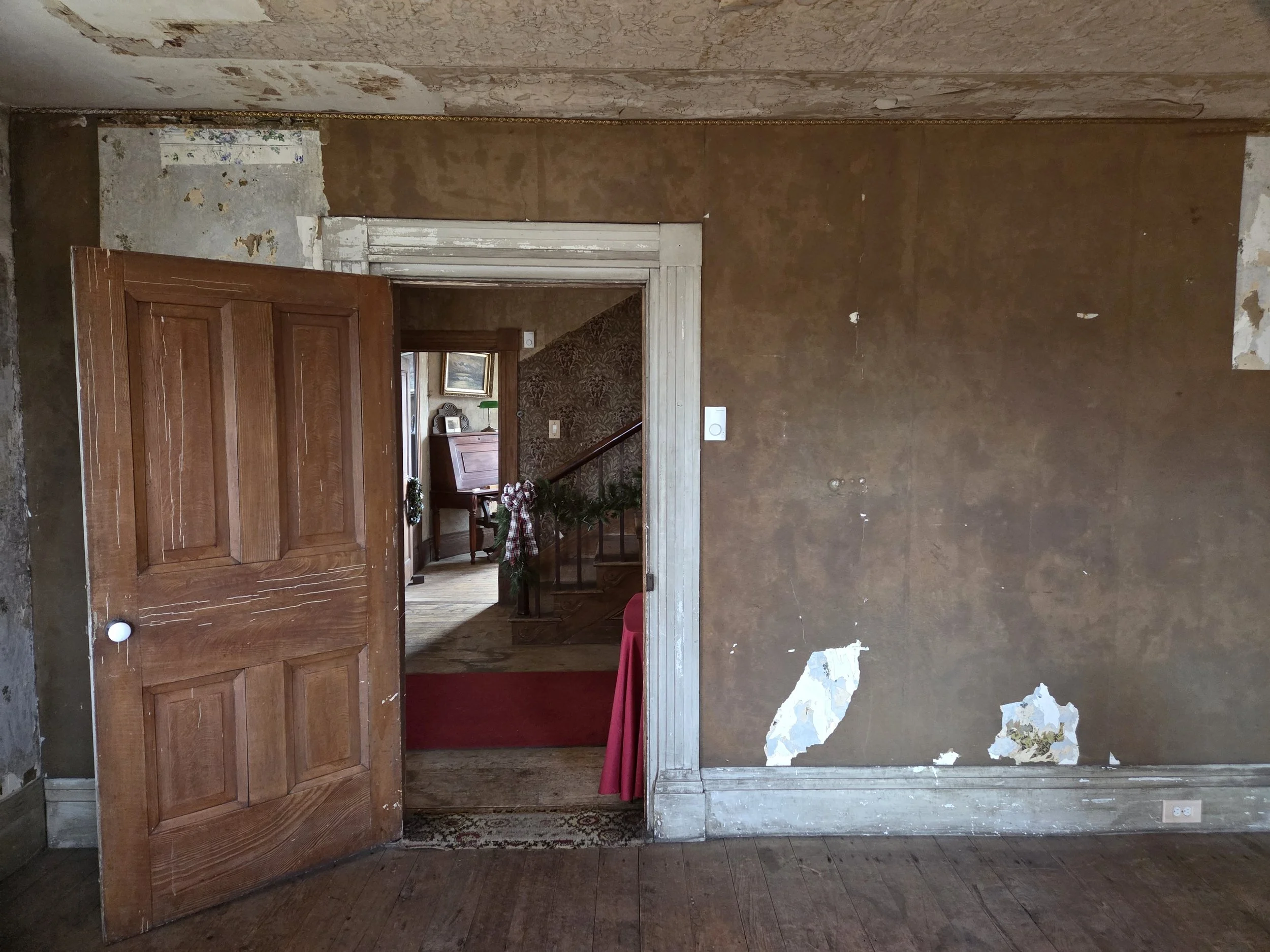 Interior of an old house with a wooden door ajar, peeling wallpaper, and a glimpse of a decorated living area with floral wallpaper, wooden furniture, and a stairway.