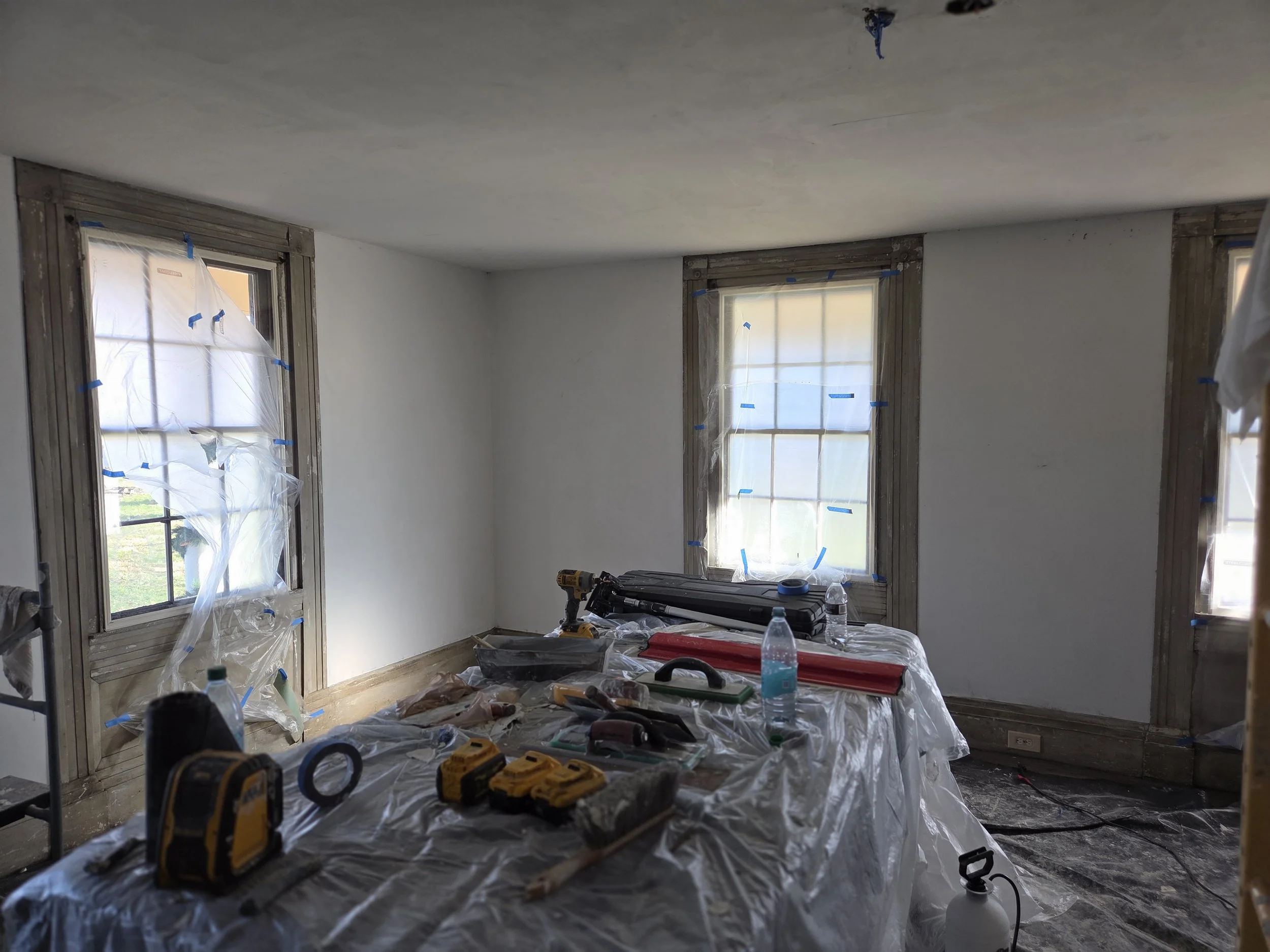 Room under renovation with window frames, tools, and materials on a covered table, plastic sheets on windows and floor.