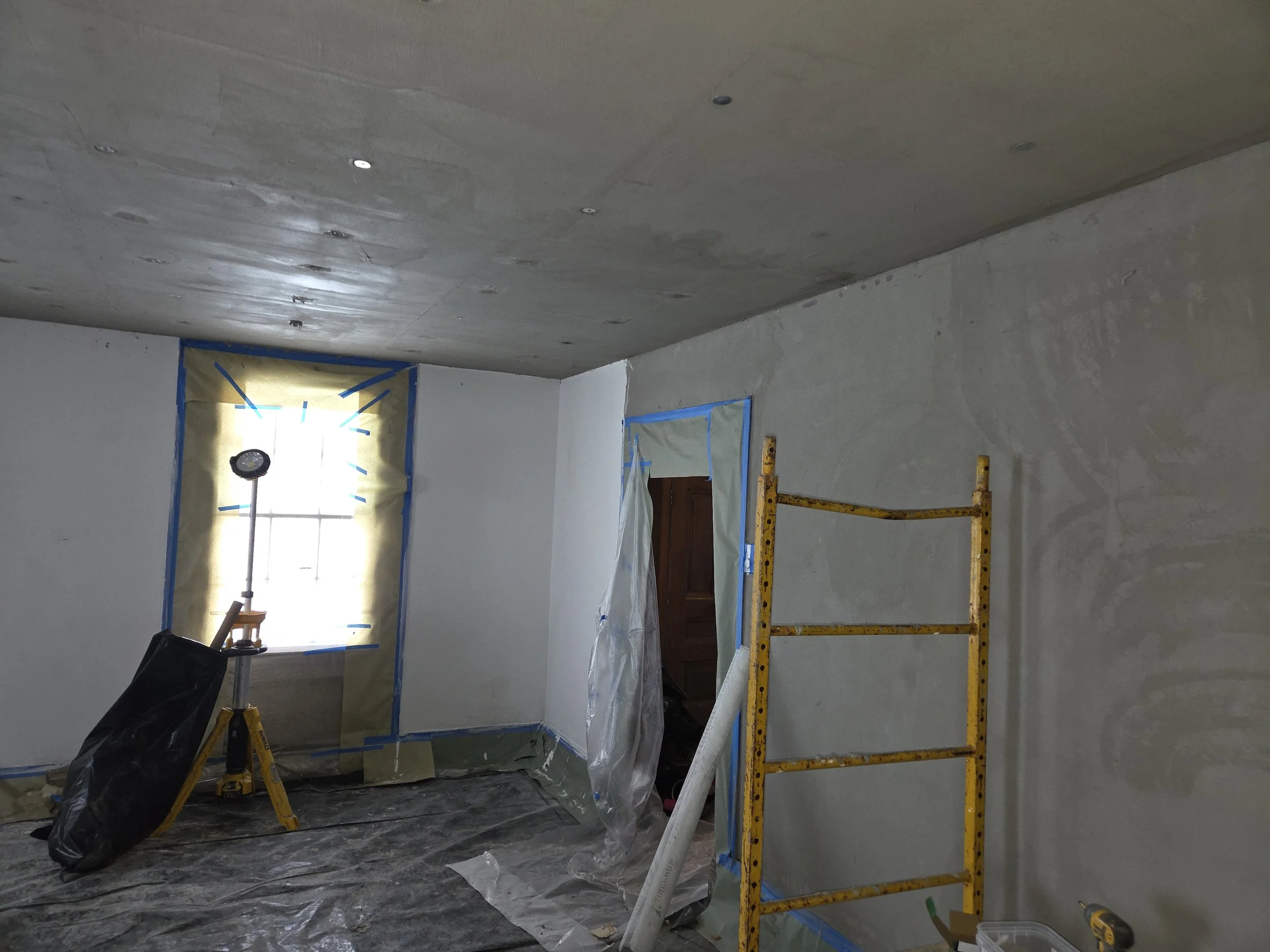 Room under renovation with taped window and doorway, construction equipment including a yellow scaffold and a work light, and plastic covering the floor.