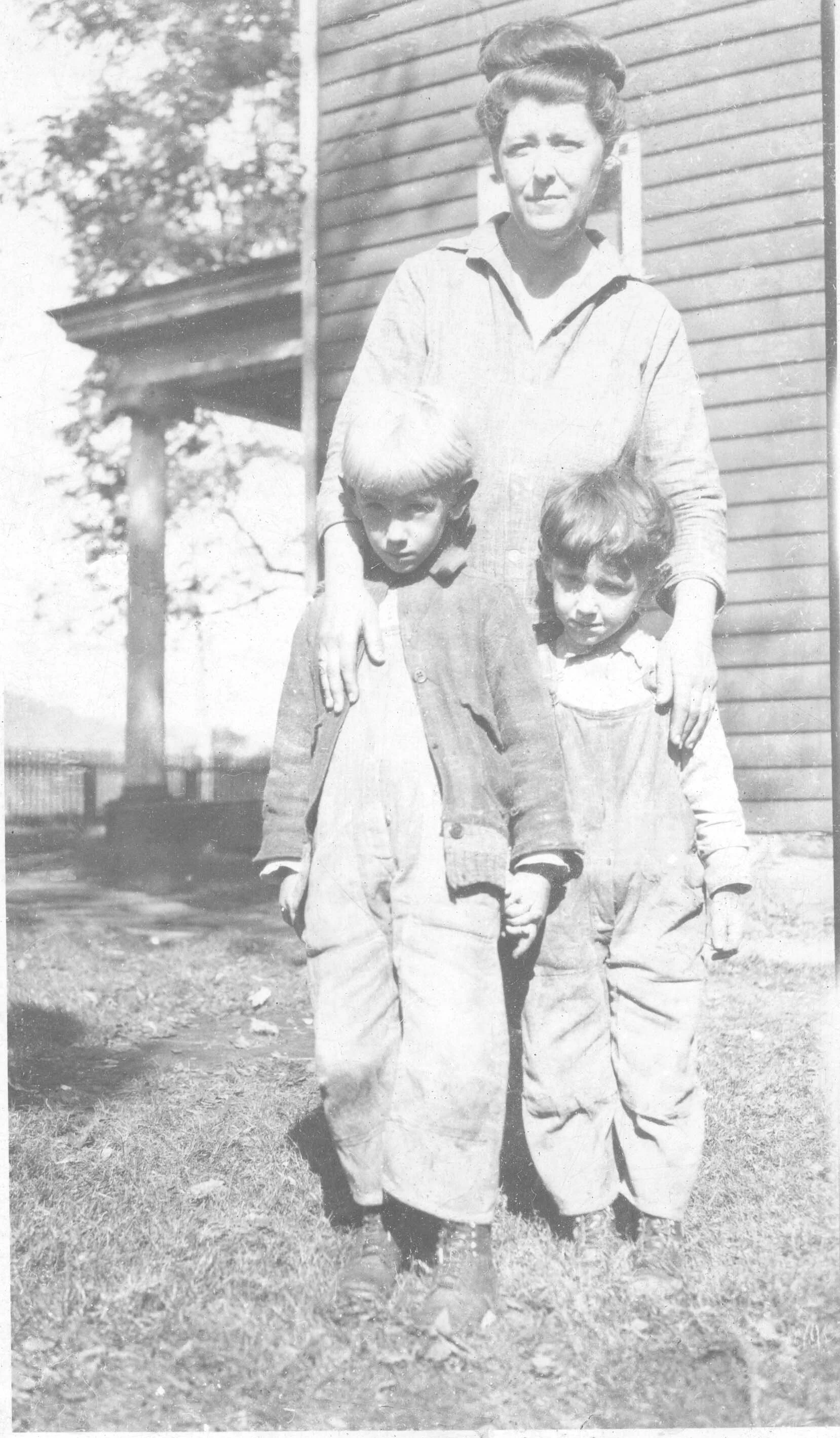 Black and white photo of Beulah with young Sam and John standing outdoors near a building with horizontal siding.