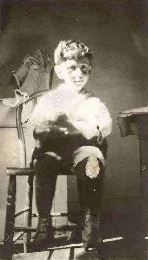 A young John Cockayne sitting on a chair with a backpack hanging on the backrest, in a vintage black-and-white photograph.