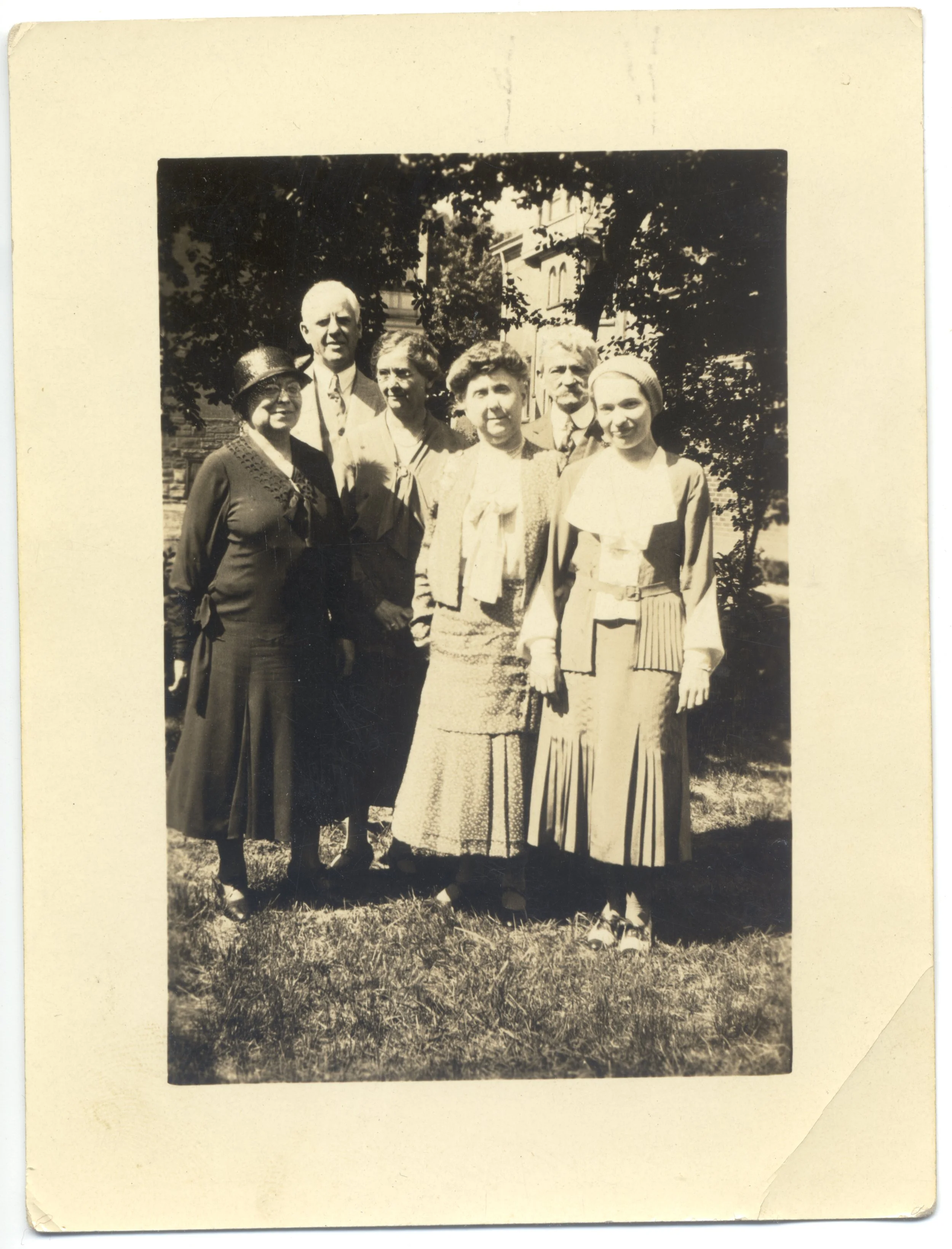 Black and white photograph of seven people standing outdoors in front of trees and a building, dressed in vintage clothing.