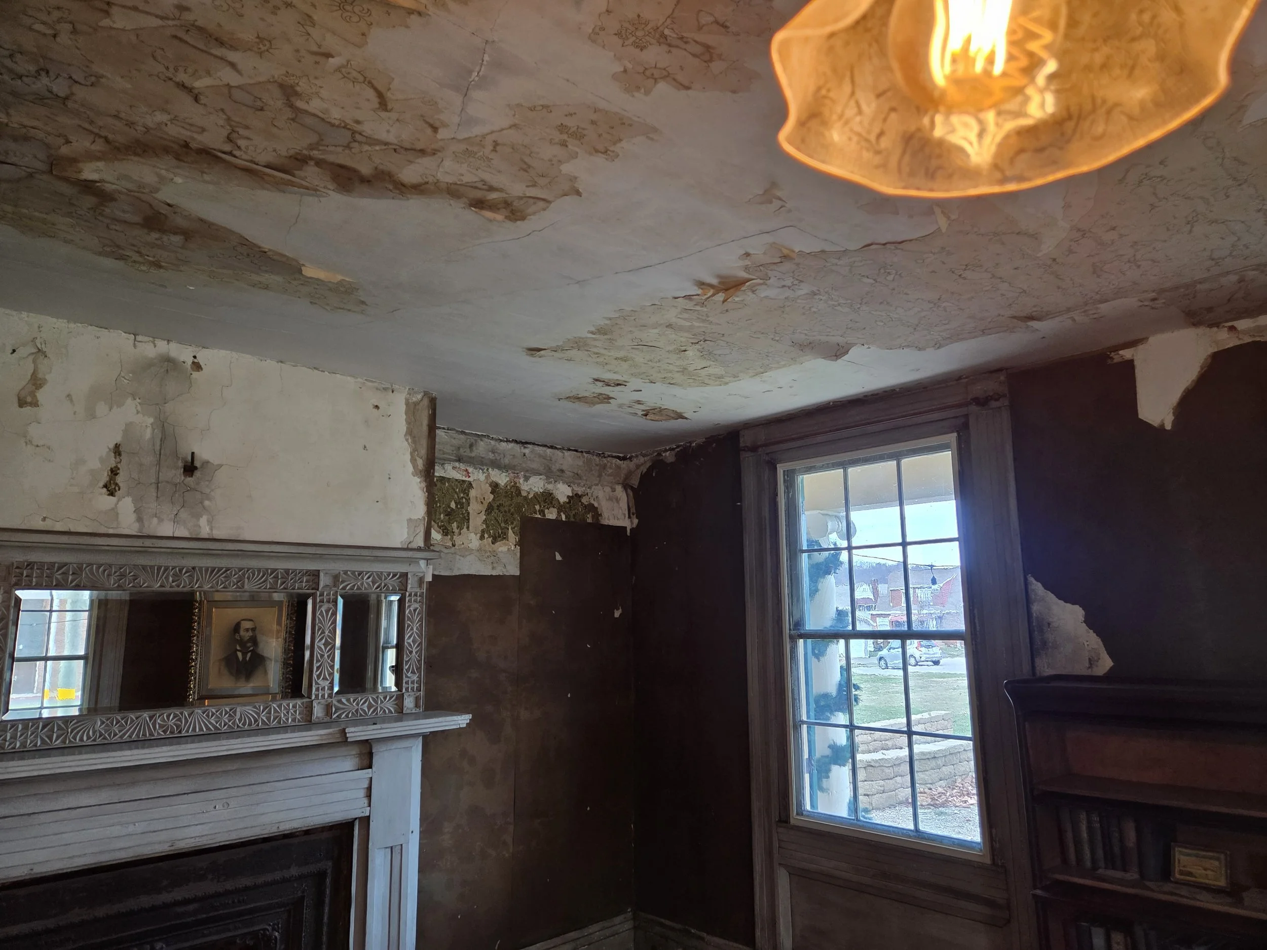 Room with peeling ceiling and walls, a window showing a street outside, a fireplace with a decorative mirror above it, and an old photograph framed on the mirror.