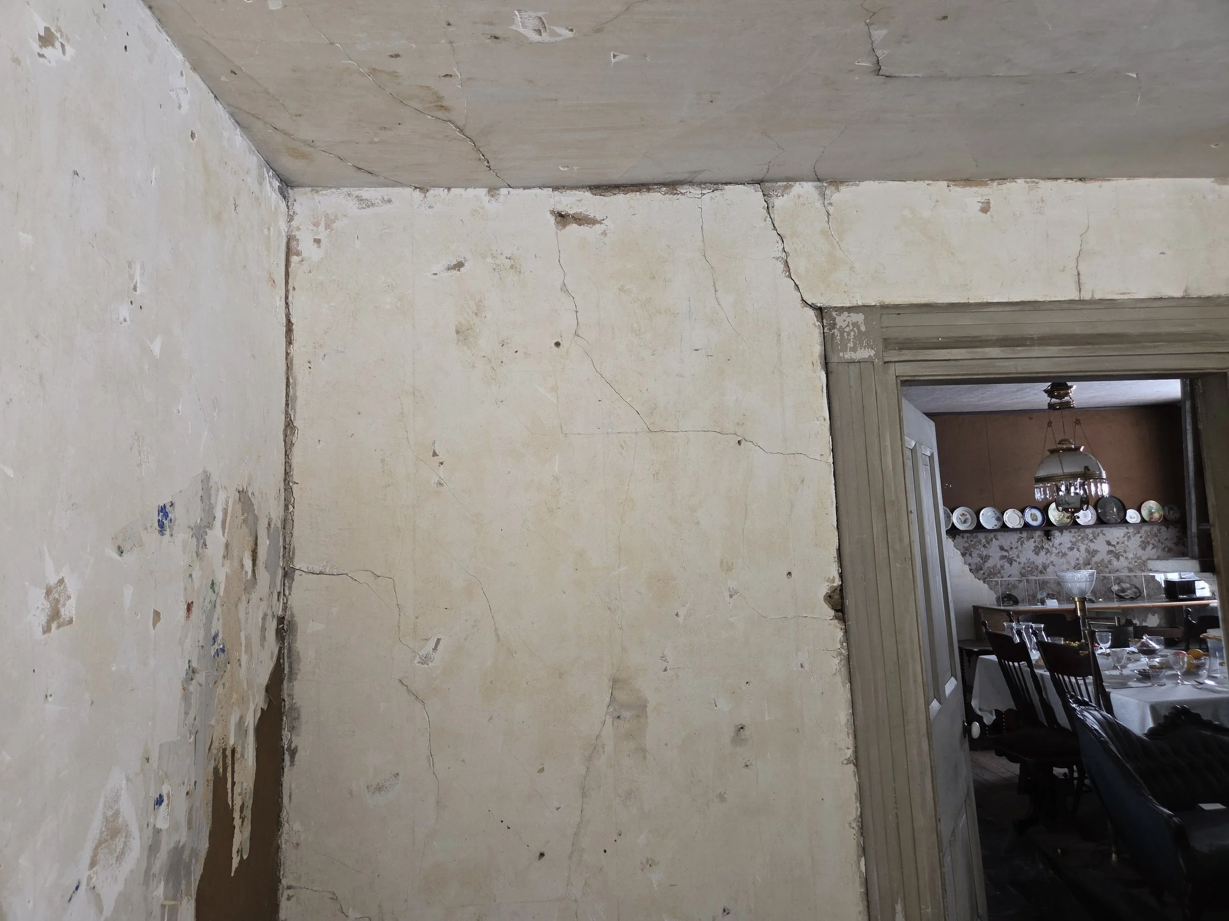 A photograph of a corner of a room with cracked and peeling paint on the walls. To the right, a doorway leads to a dining area with a table set for a meal, with plates on the wall and a hanging light fixture.