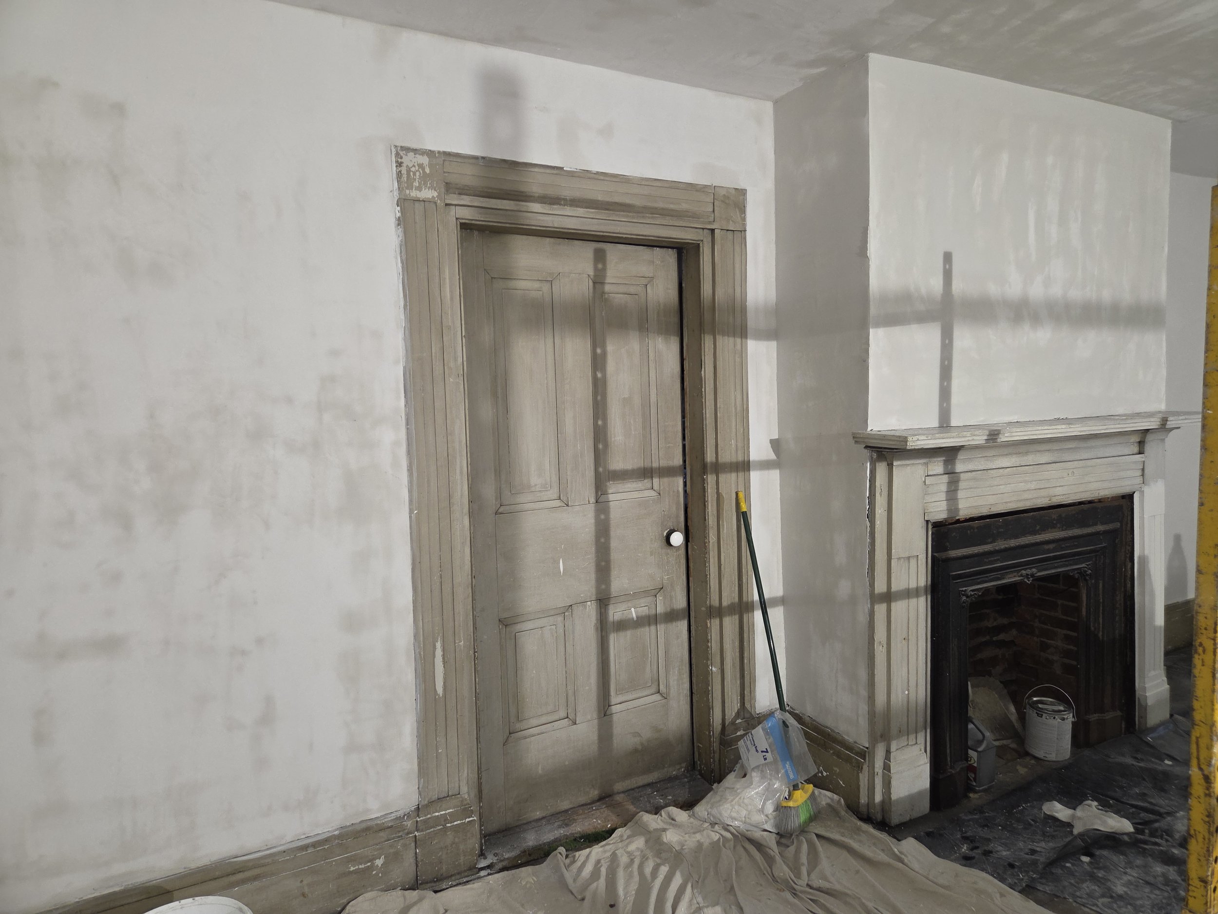 Interior wall undergoing renovation with a wooden door frame and a fireplace, construction tools and materials on the floor.