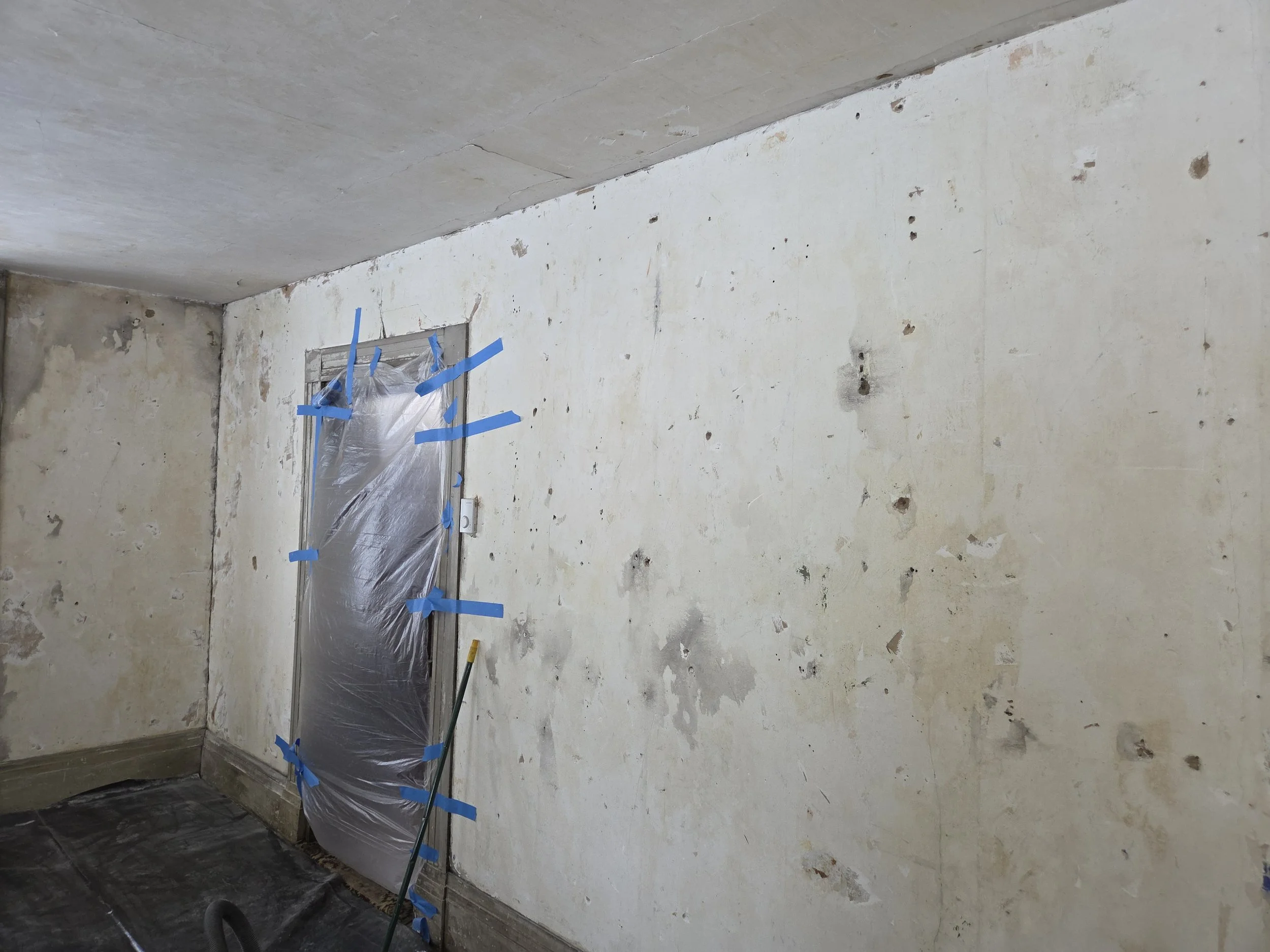A partially renovated room with a wall marked with several spots and a door covered with plastic secured with blue painter's tape.