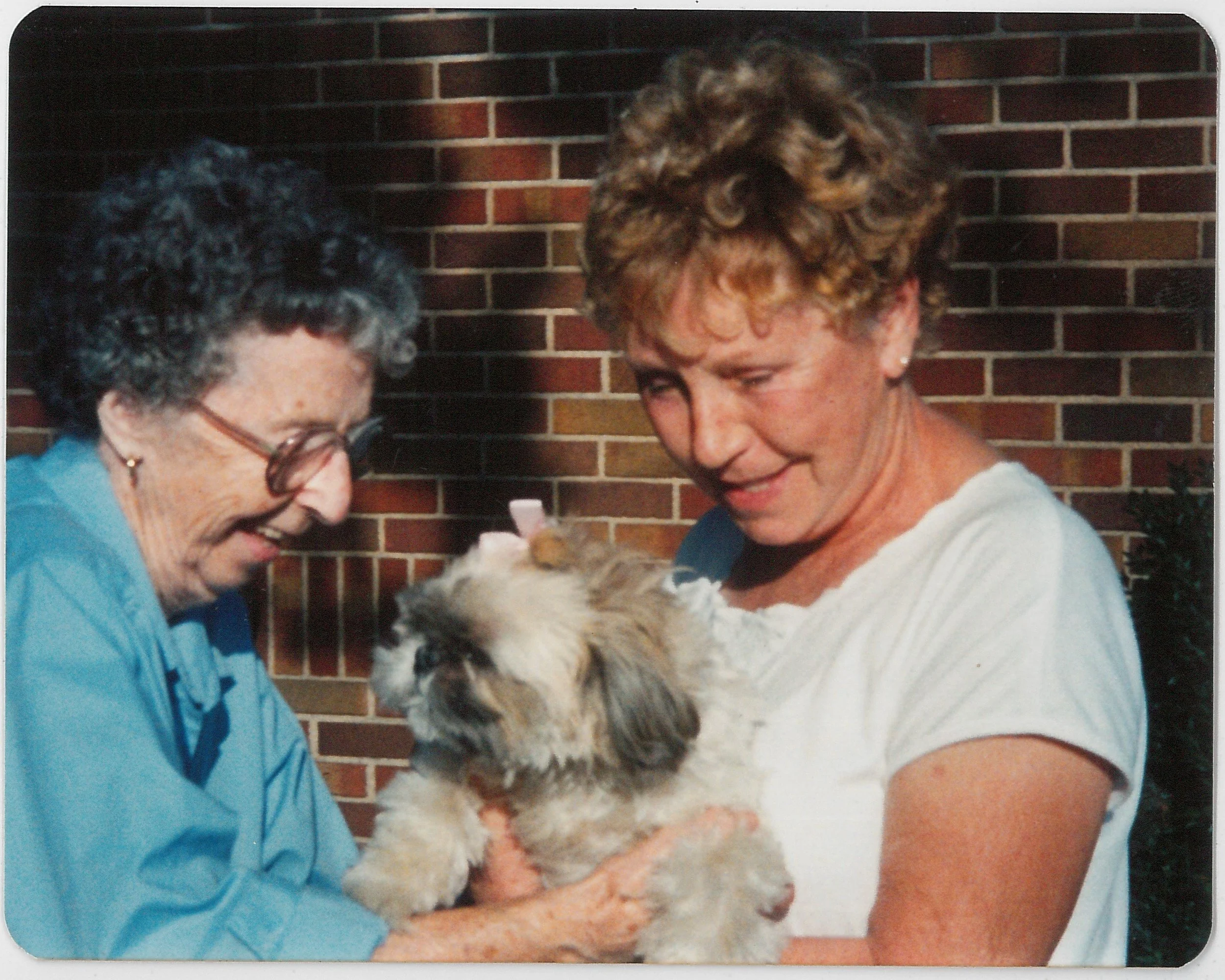 Two women smiling and holding a fluffy dog together outside in front of a brick wall.