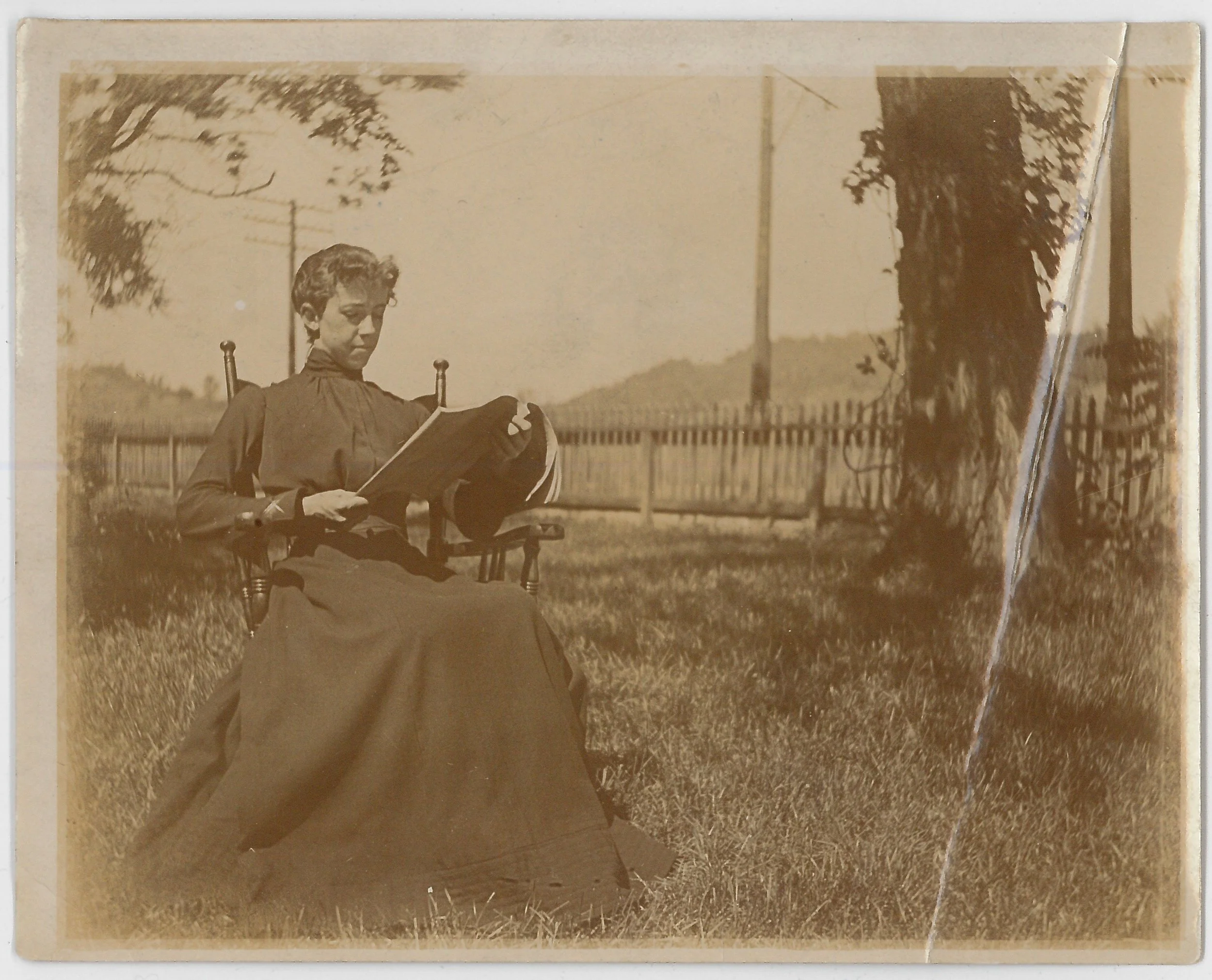 A woman in a long dark dress sitting on a wooden chair outdoors, reading a book, with trees and a fence in the background.