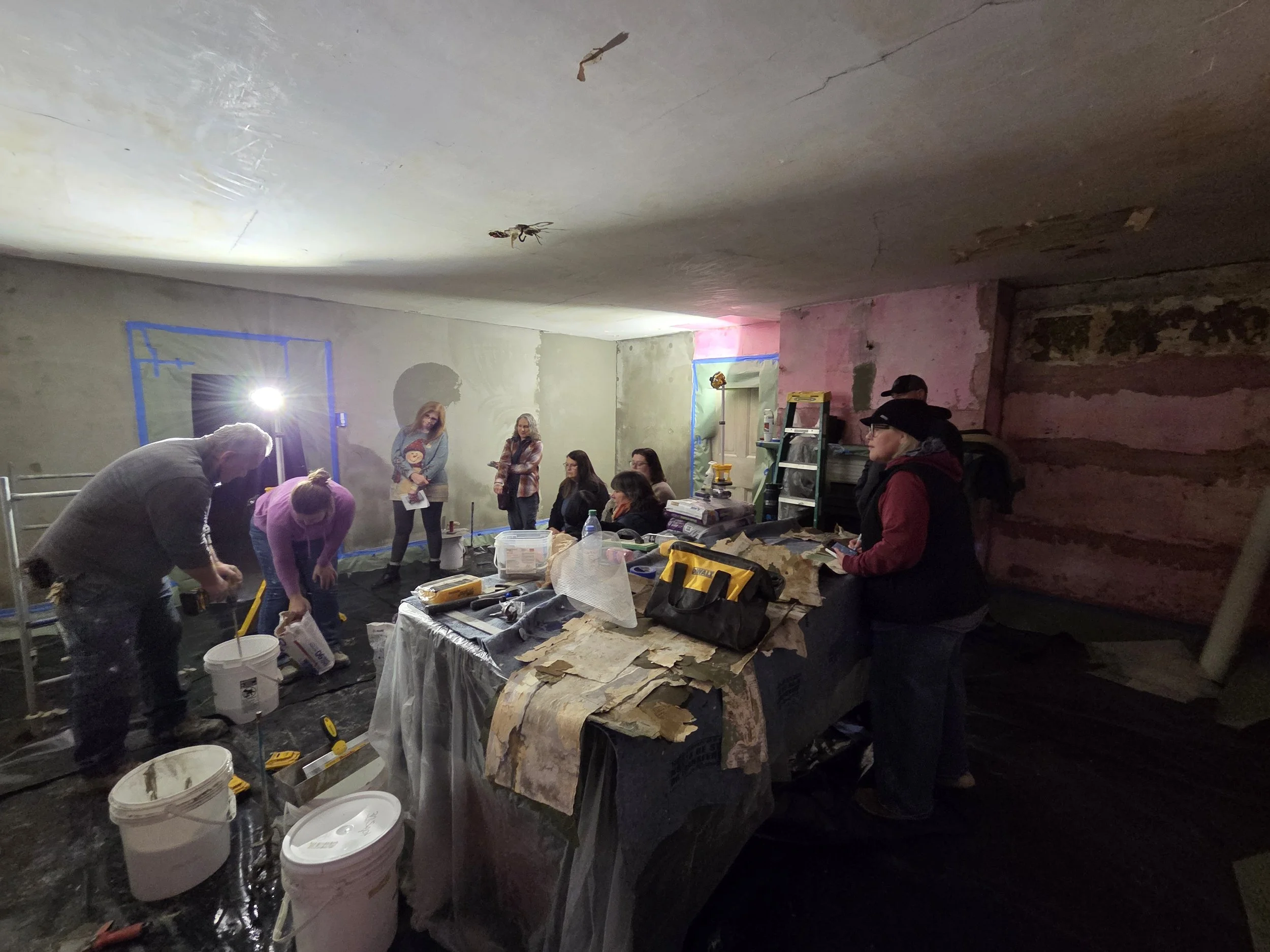 People working on a construction or renovation project in an indoor space with unfinished walls and ceiling, using various tools and materials.