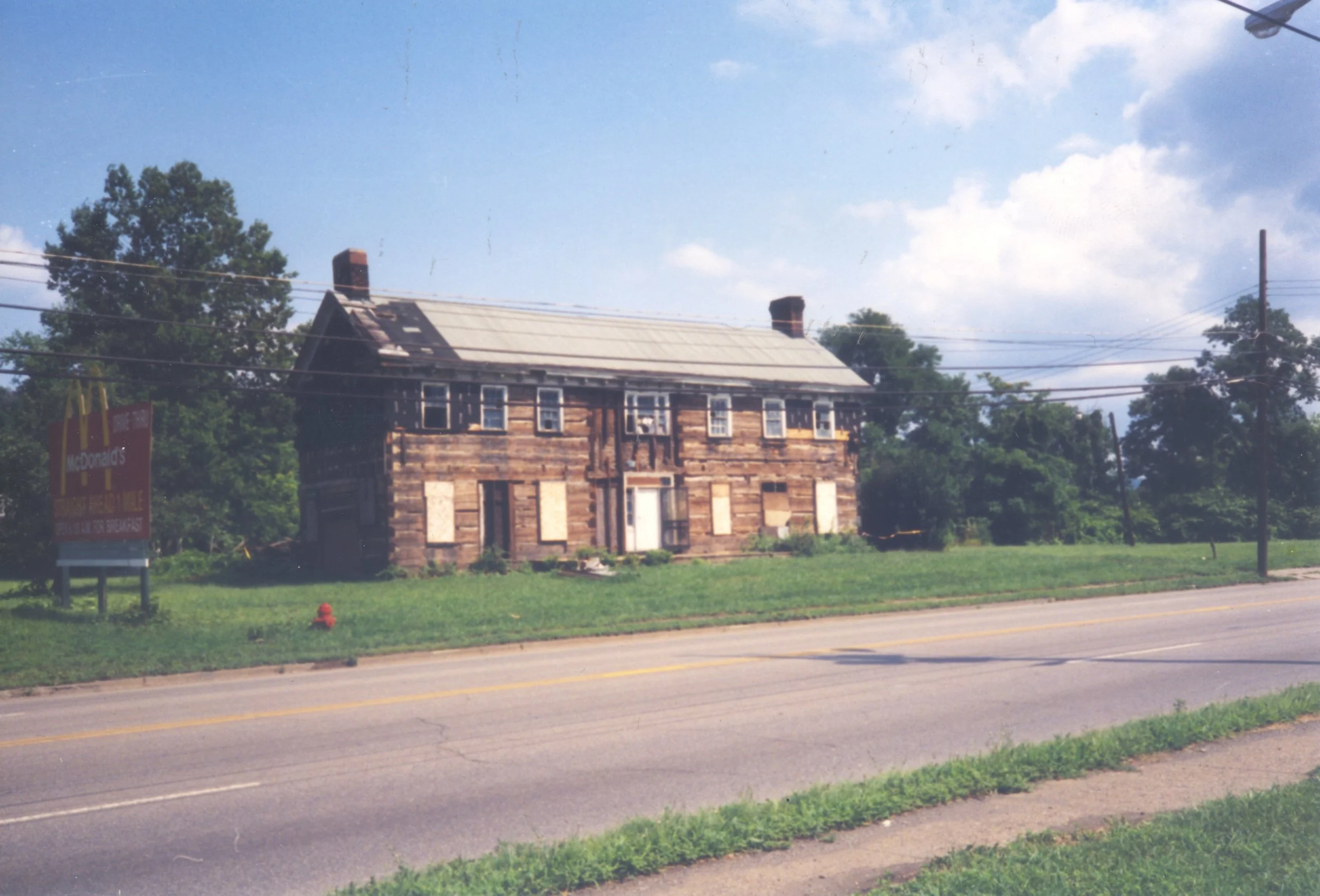 An abandoned two-story brick house with boarded-up windows, situated along a grassy roadside, with a McDonald's sign visible on the left. The house appears weathered, with a damaged roof, and is surrounded by trees and utility lines under a partly cloudy sky.