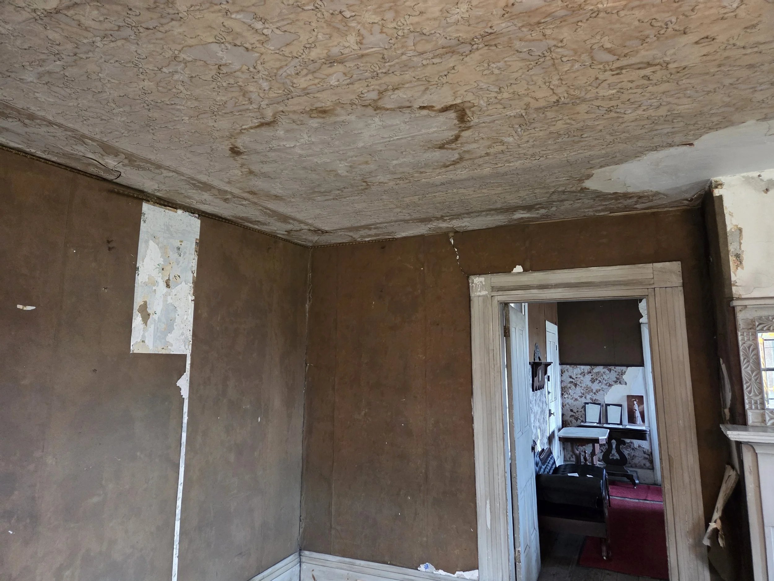 A room with peeling wallpaper and deteriorating ceiling, visible cracks, and damaged trim surrounding a doorway leading to another room with floral wallpaper, framed photos, and furniture.